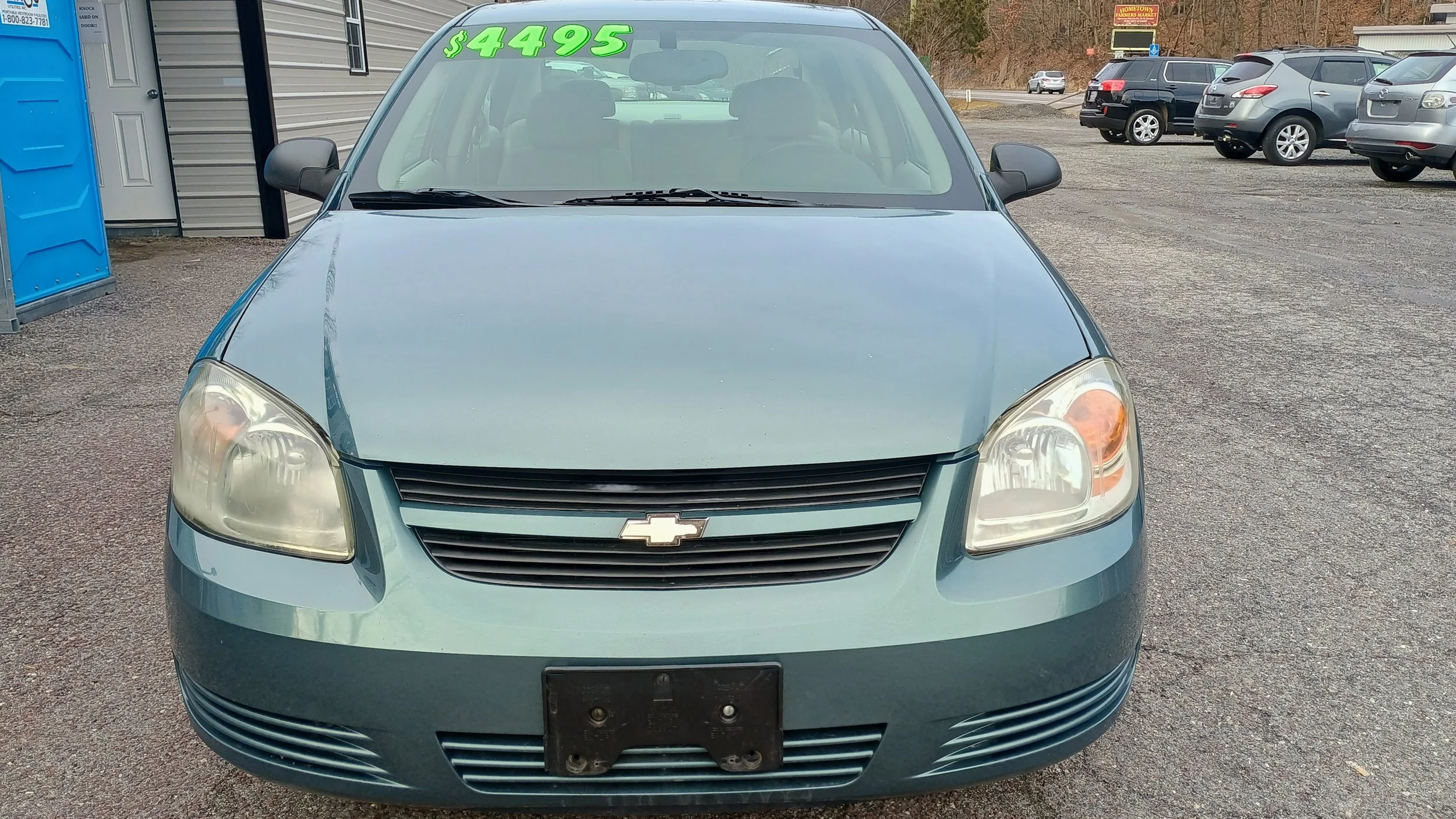 Front view of a light blue Chevrolet sedan displaying a green price sign of $4495 on the windshield, parked in a lot with other vehicles and a portable toilet nearby.
