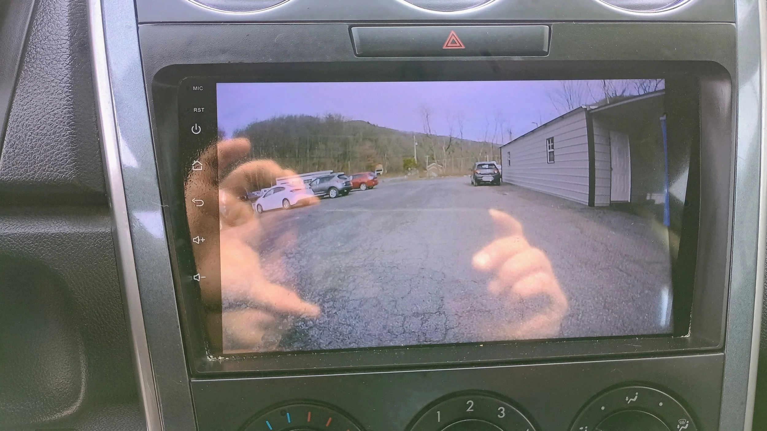 Car dashboard screen displaying a rearview camera image of a parking lot with several parked cars and a building on the right side.
