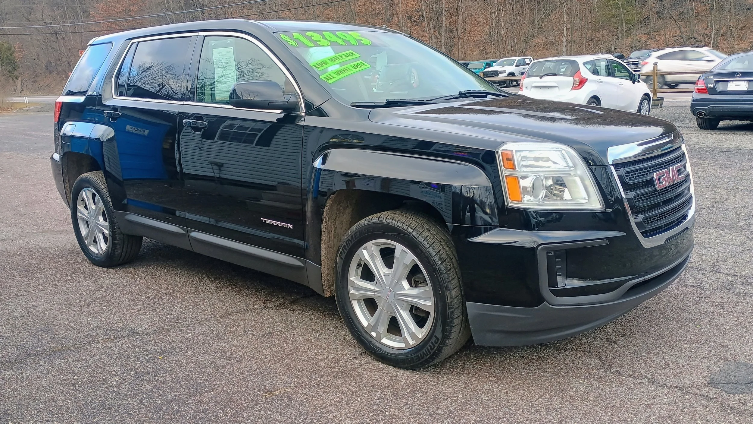 Black GMC Terrain SUV with green price and features signs on the windshield parked on a gravel lot with other cars in the background.
