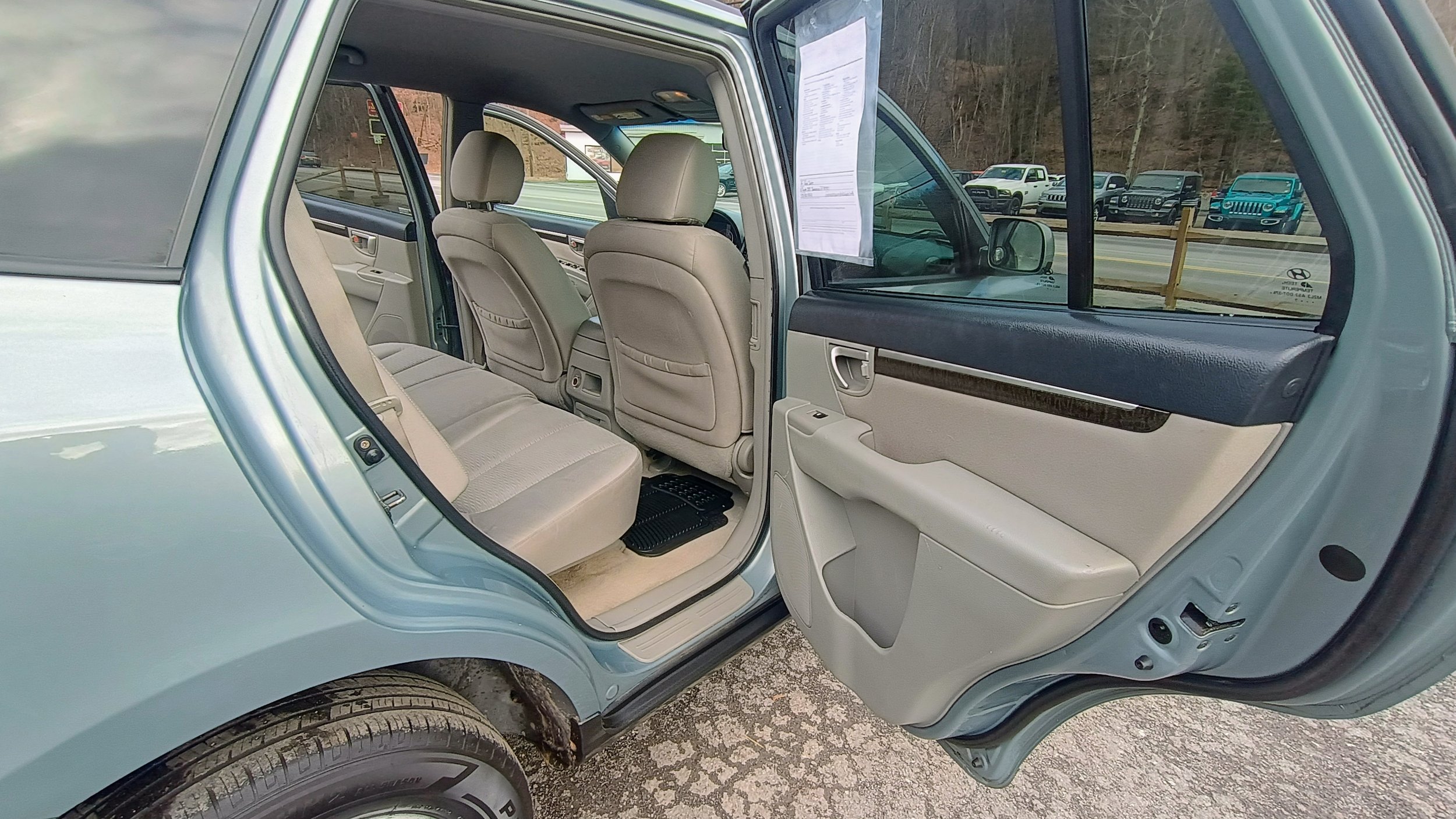 The image shows the interior of a light-colored SUV viewed with the rear door open. The back seats and front seats are visible, with beige upholstery. There is a paper document attached to the inside of the rear window. In the background, there are parked cars, a road, and trees.