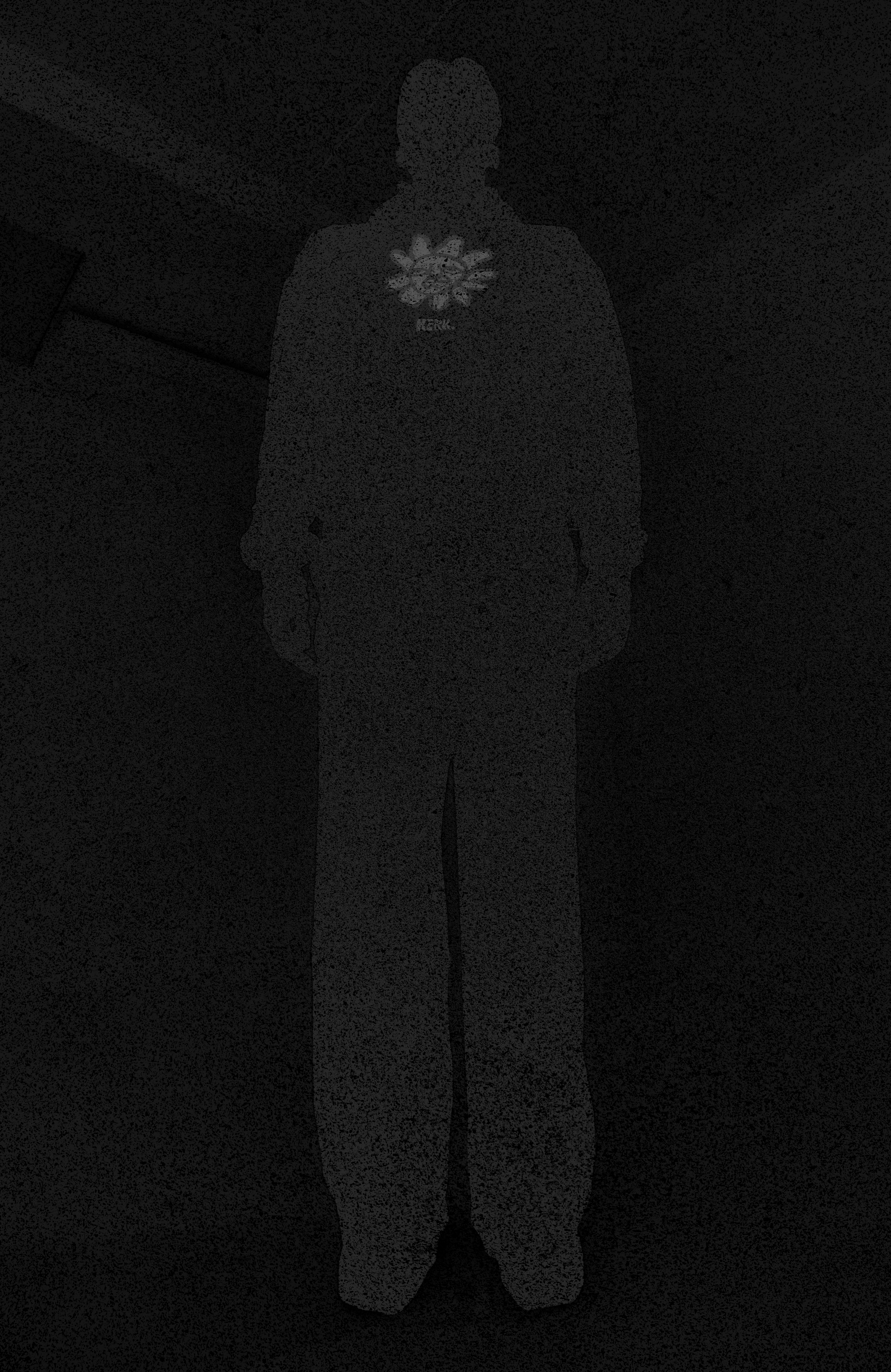 A person standing in a dark room, facing forward, wearing a jacket with a large floral design on the chest.