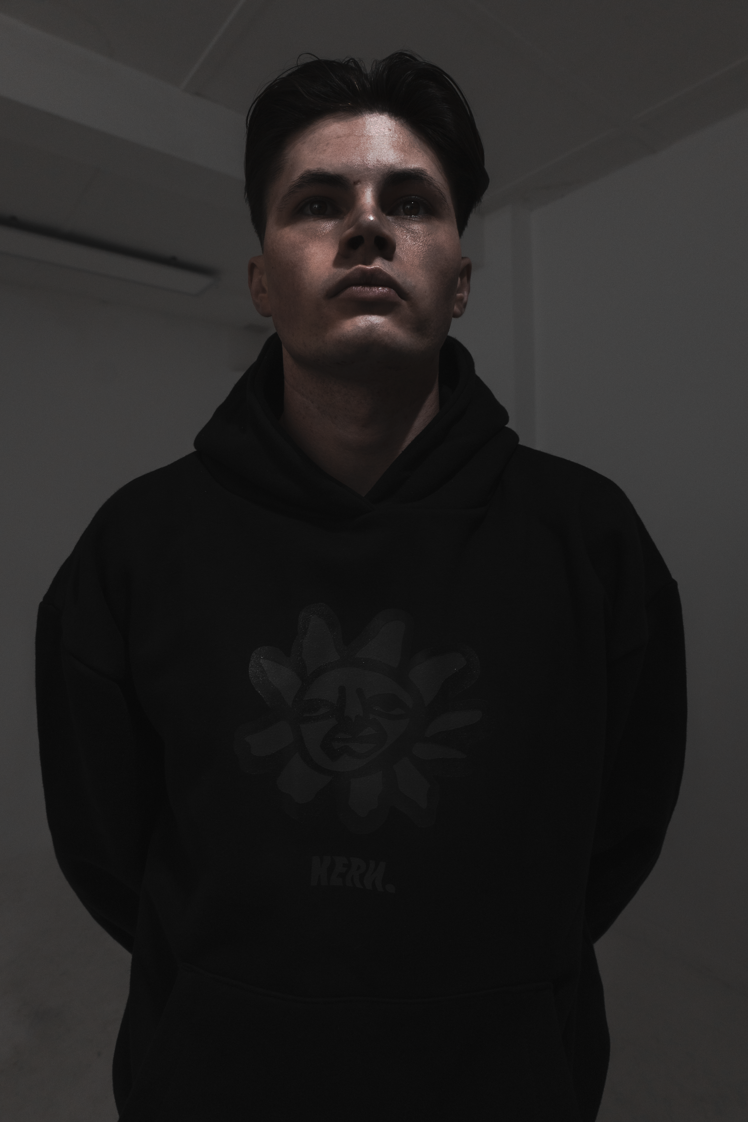 A young man with dark hair wearing a black hoodie with a graphic design stands in a dimly lit room.