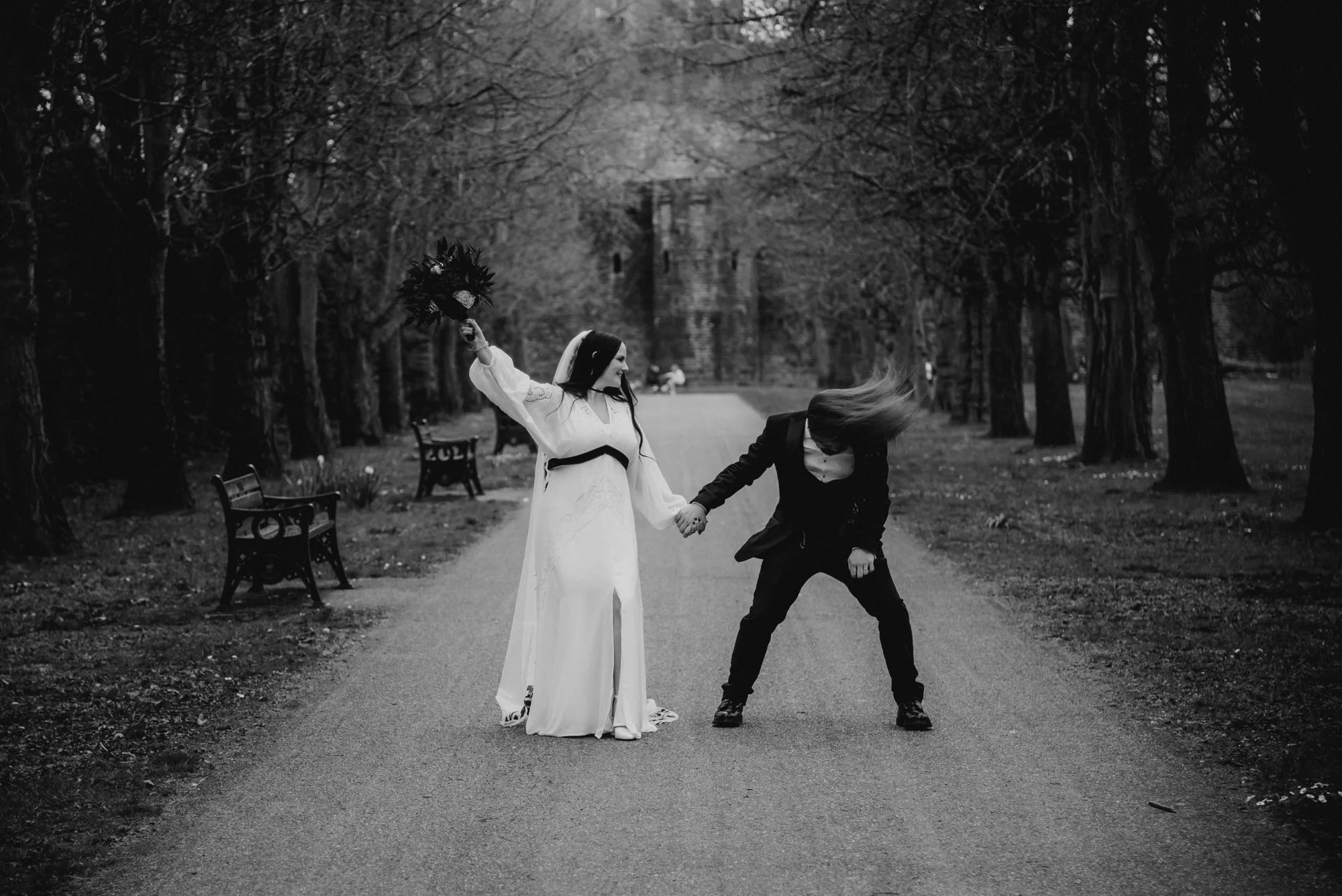 A bride and groom are holding hands on a tree-lined park path, with a castle in the background. The bride is holding a bouquet and wearing a long white dress, while the groom is dressed in a black suit. The scene appears to be lively and joyful.