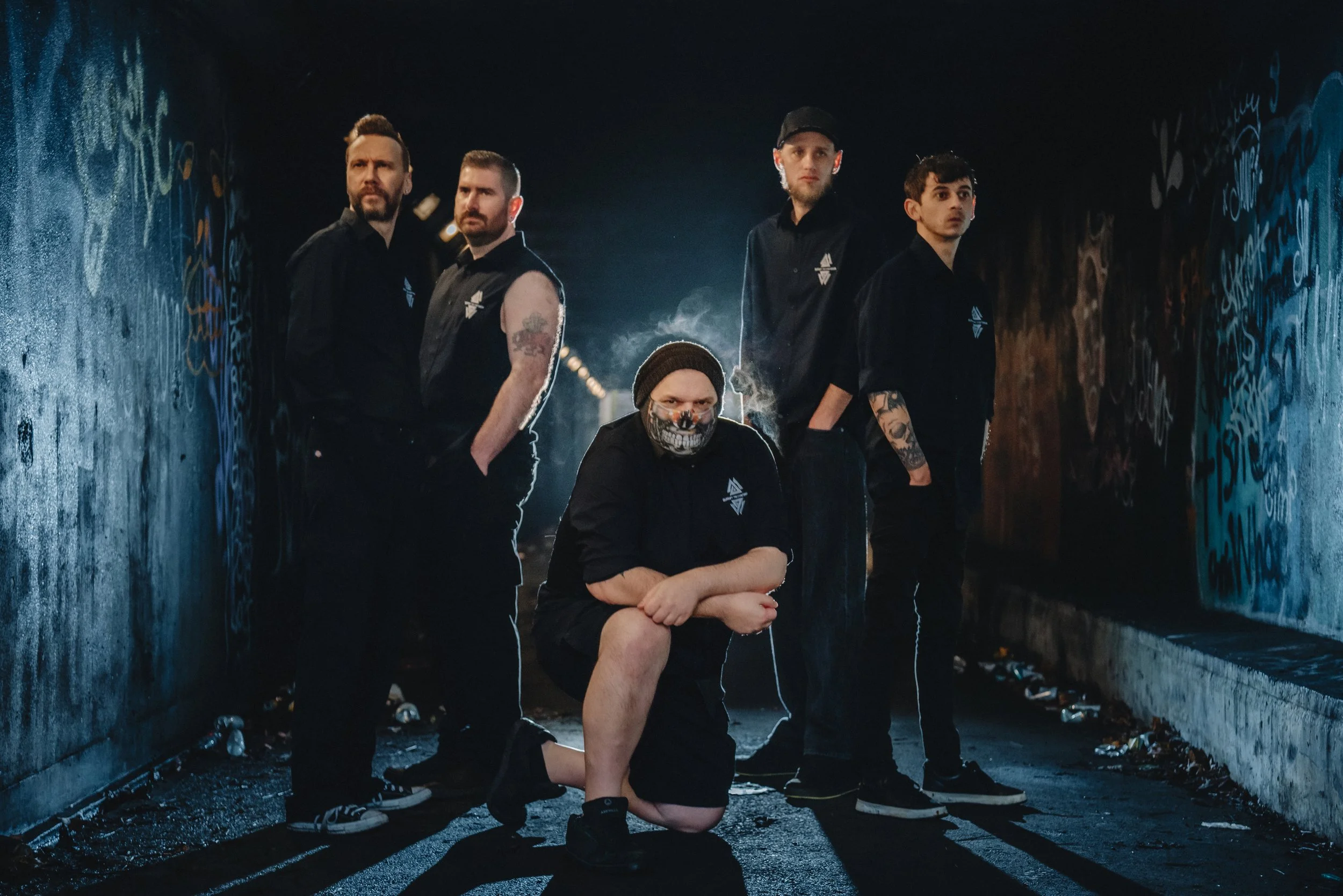 Five men standing in a dark graffiti-covered tunnel, with one crouching in front wearing a skull mask, all dressed in black.