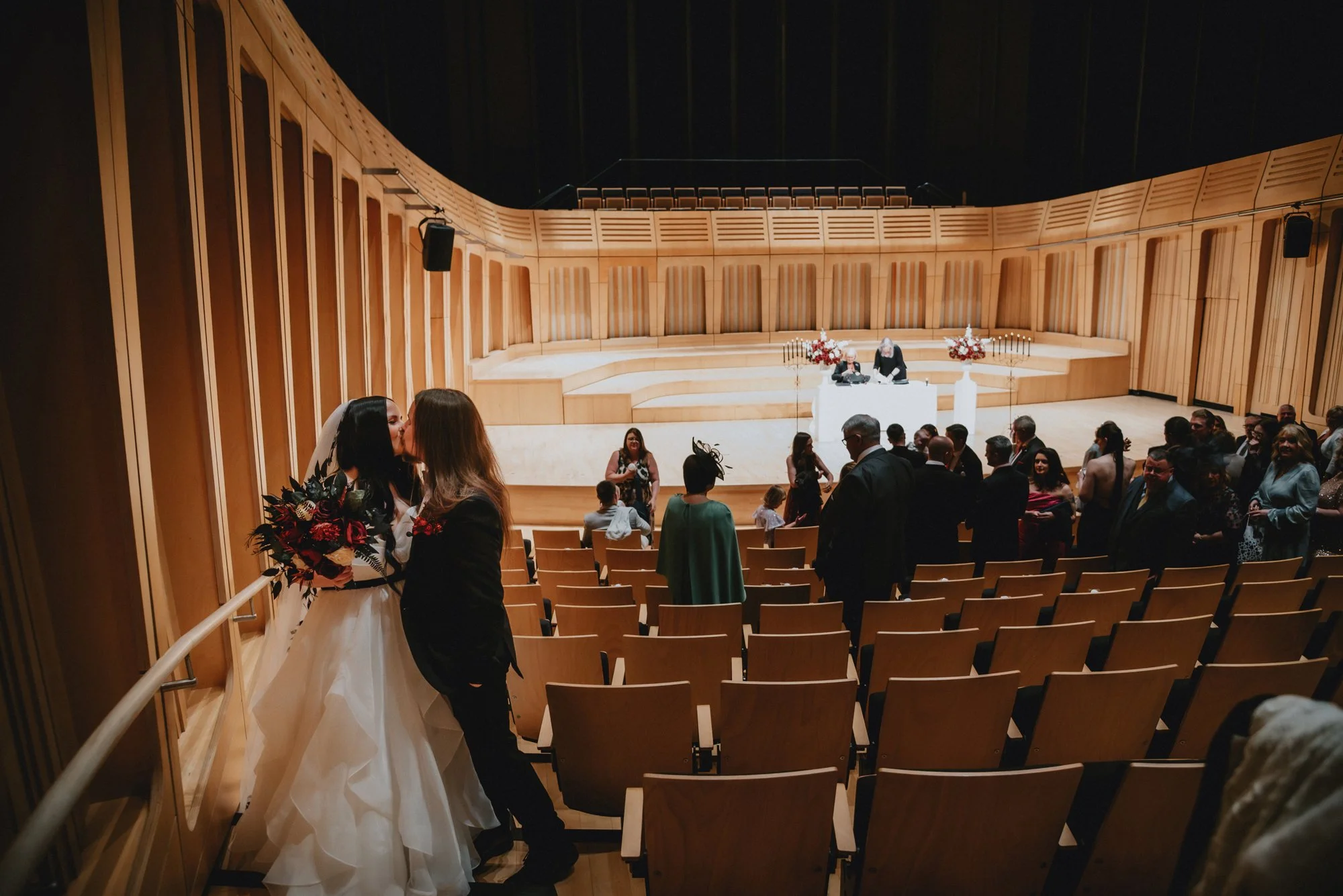 Two women, one in a wedding dress and the other in a black dress, sharing a kiss at a wedding ceremony inside a concert hall or auditorium with wooden walls and seating. Guests are standing and talking near the stage, which is decorated with flowers 