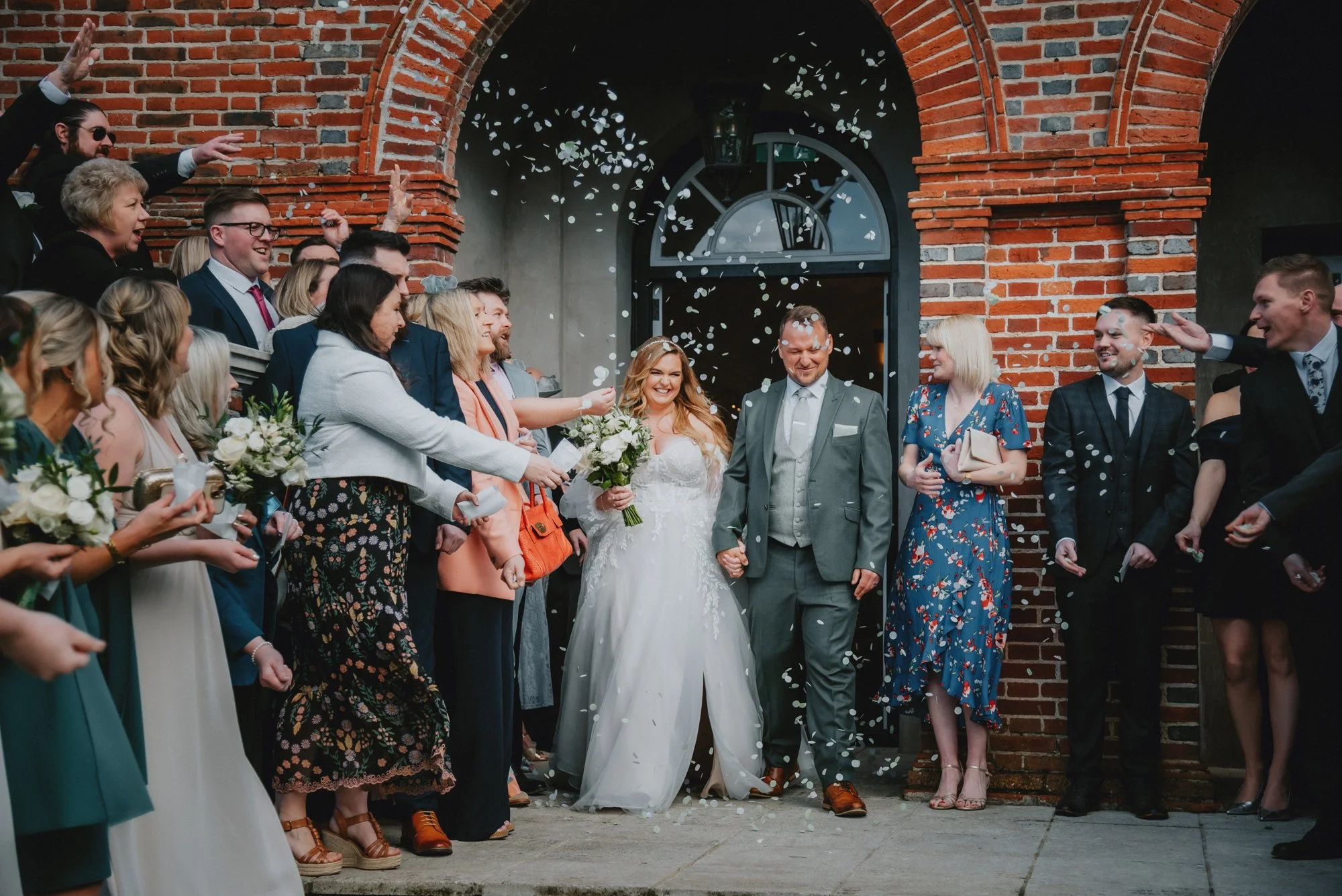 A bride and groom walking out of a wedding venue, holding hands, surrounded by wedding guests showering them with confetti.