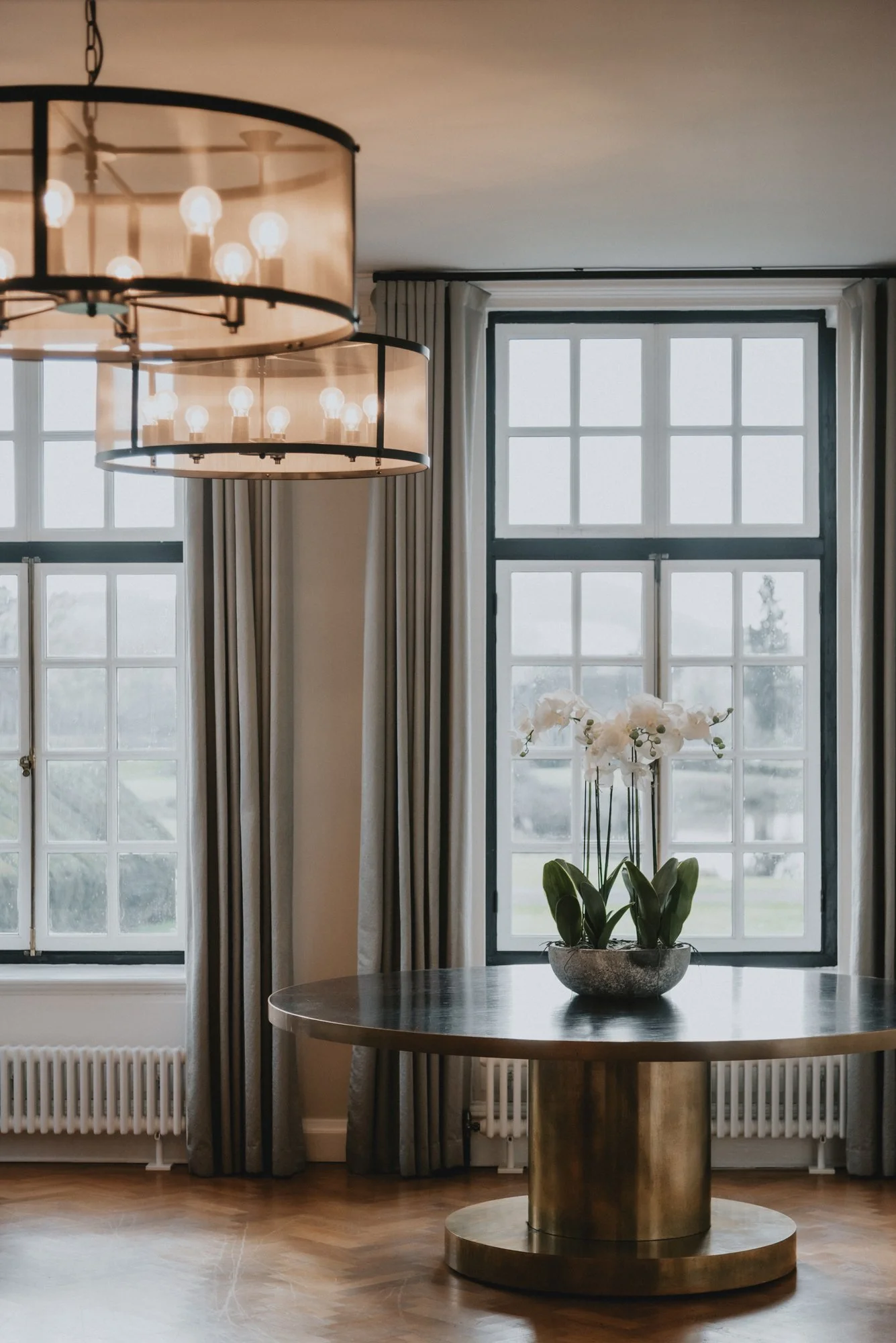Interior of a room featuring a round wooden table with a potted orchid, large windows with divided panes, curtains, and a chandelier with exposed bulbs.
