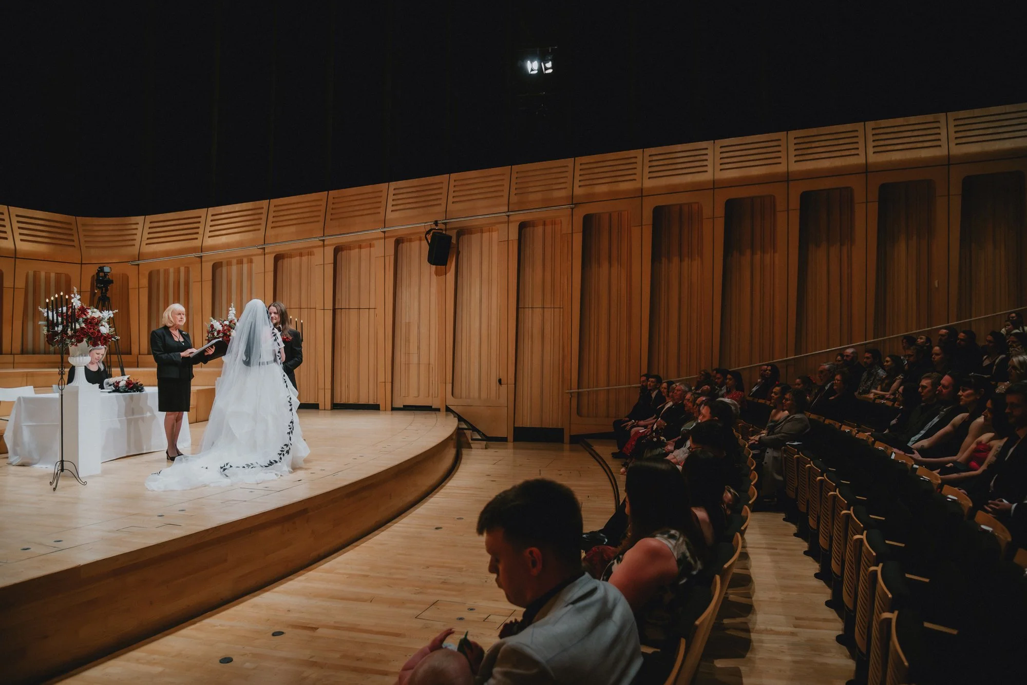 A wedding ceremony taking place inside a wooden auditorium. A bride in a white gown and veil is standing with an officiant and another woman. Guests are seated facing the stage, watching the ceremony.