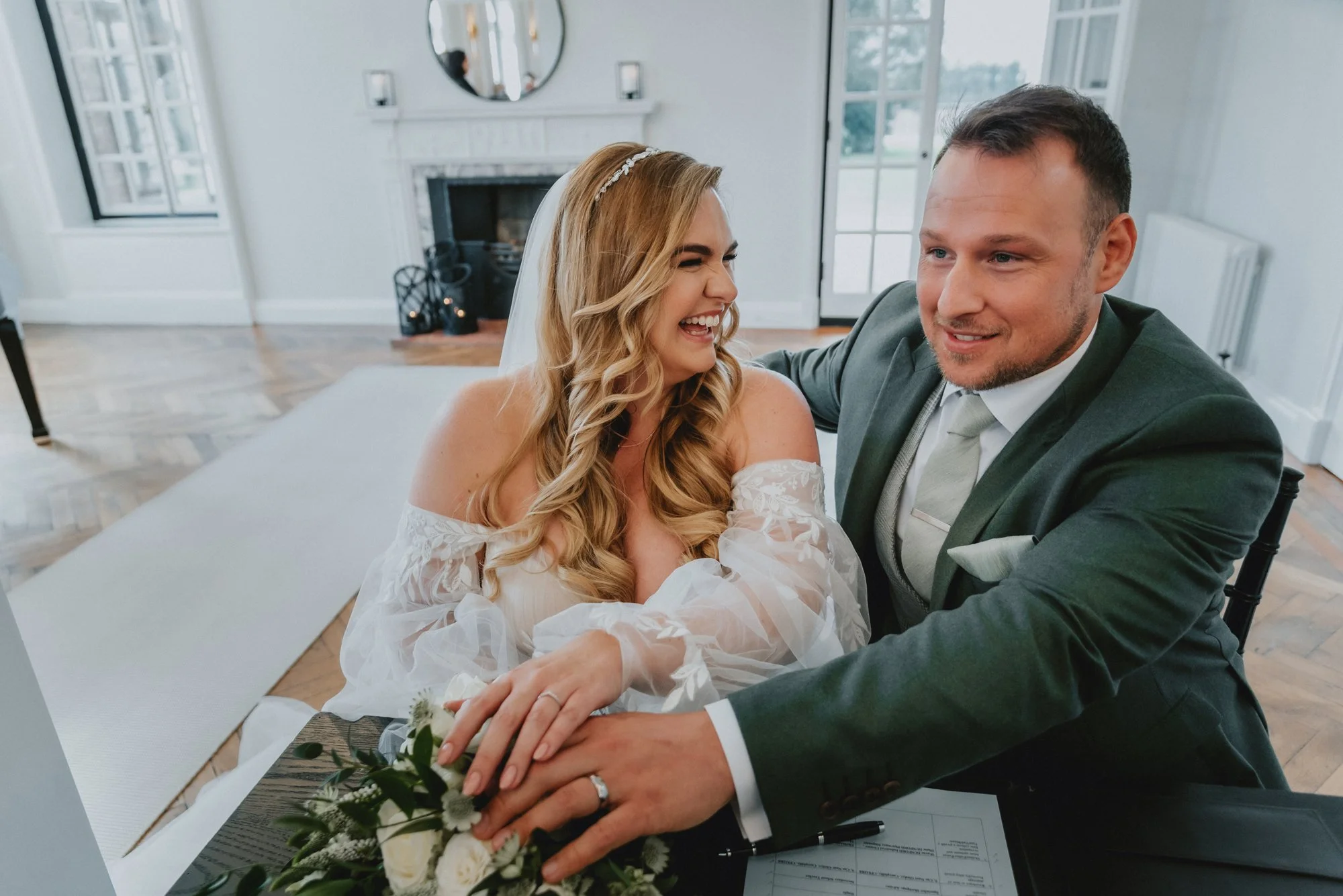 A newlywed couple sits at a table, smiling and holding hands, during their wedding ceremony in a bright, elegant room with large windows, a fireplace, and a mirror above it.