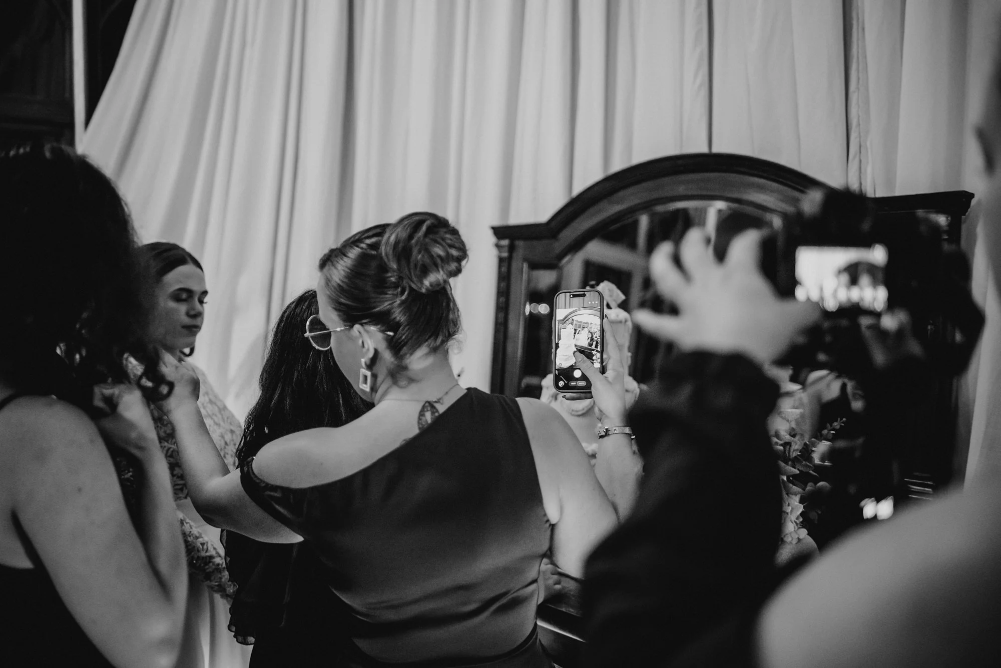Women taking pictures of a woman, possibly at a wedding or special event, in front of a mirror with a curtain backdrop.
