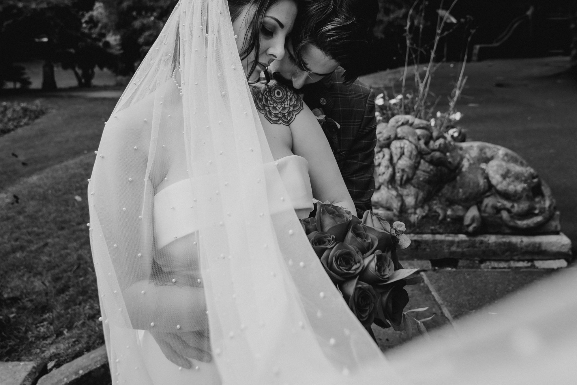 A bride and groom share an intimate moment outdoors during a wedding, with the bride holding a bouquet of roses, wearing a veil and dress, and the groom embracing her from behind.