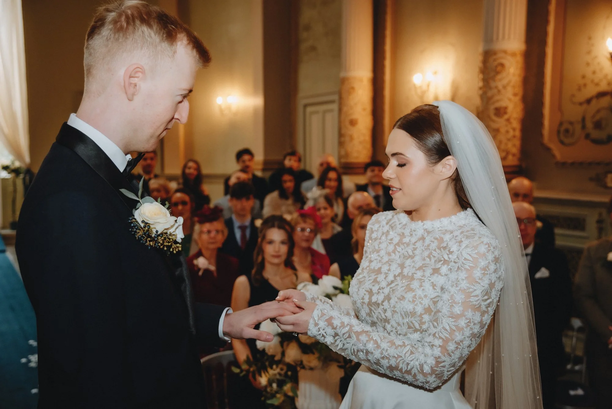 A bride and groom exchanging rings during their wedding ceremony in an elegant decorated hall, with seated guests watching.
