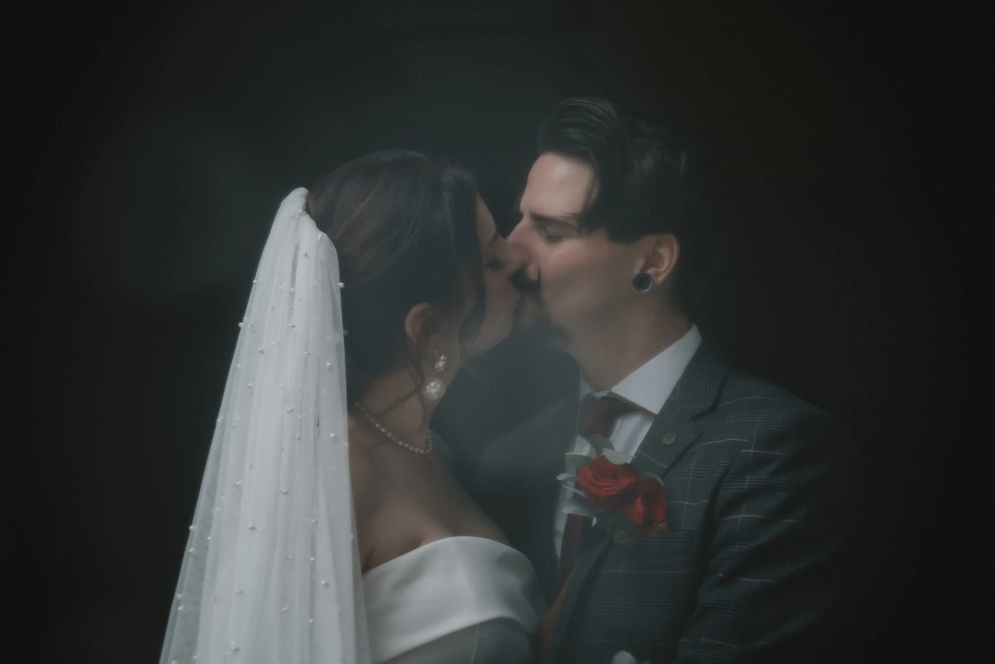 A bride and groom share a kiss, with the bride wearing a white veil and wedding dress, and the groom dressed in a plaid suit with a red boutonniere, against a dark background.