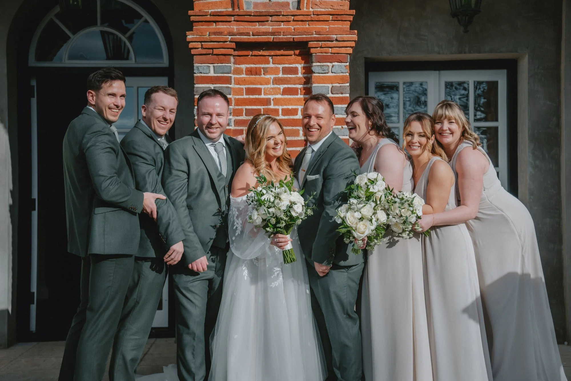 Bride and groom with wedding party smiling outside in front of brick wall and windows
