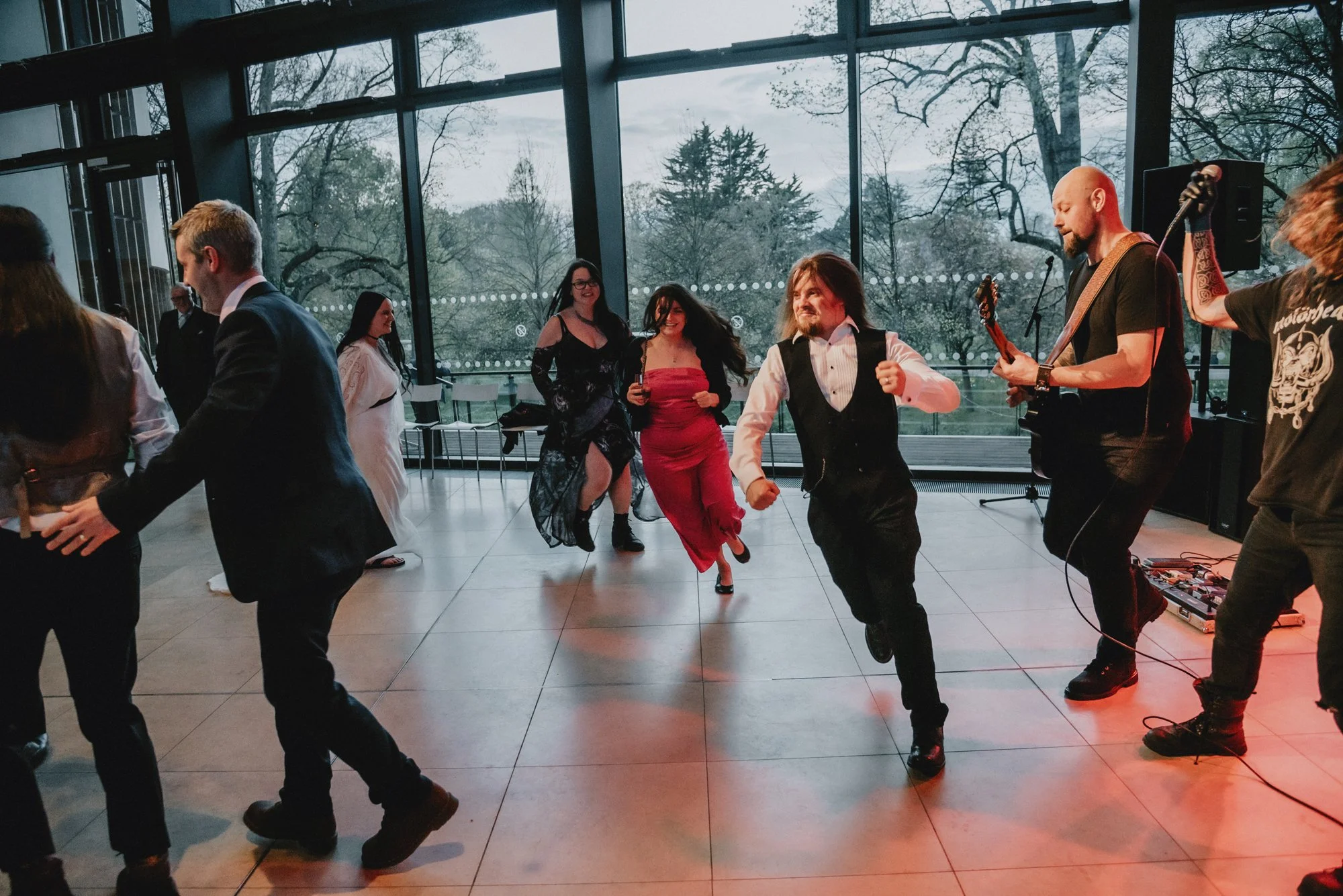 People dancing and a live band performing at an indoor event with large windows and trees outside.