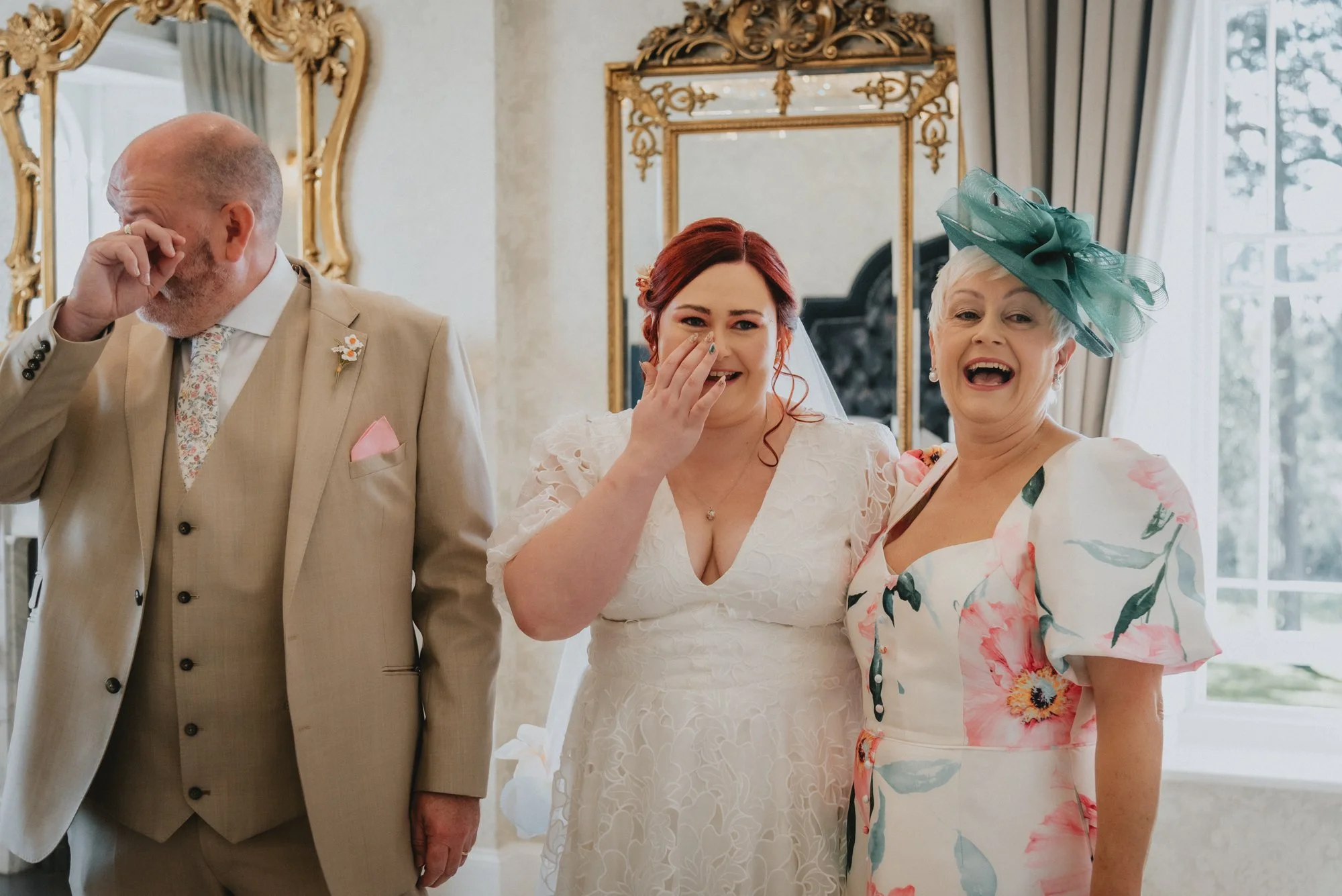 Three people at a wedding reception are emotionally reacting, with one woman in the center wiping tears, another woman on the right wearing a floral dress and hat smiling, and a man on the left in a beige suit with his hand to his face.