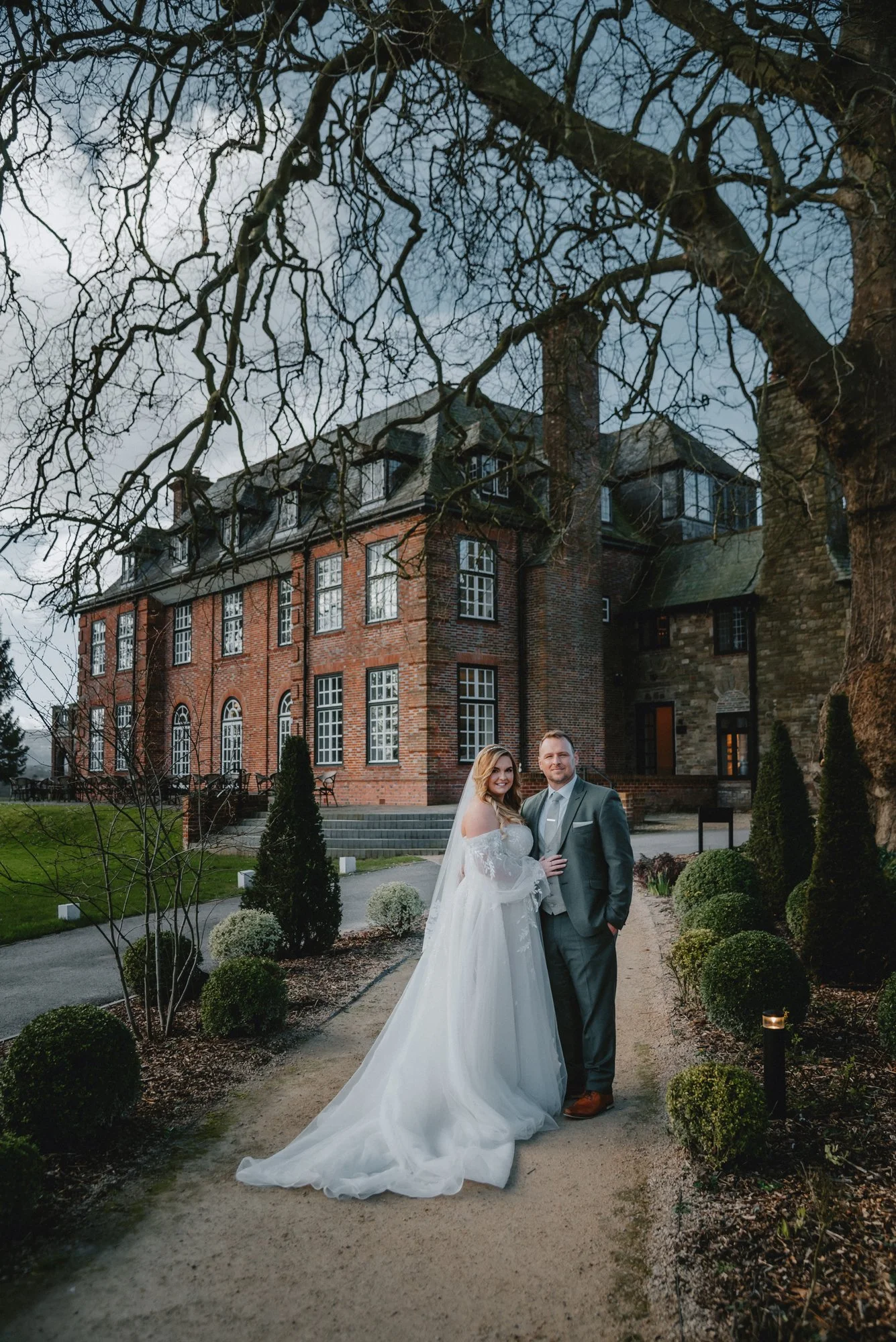 A bride and groom in wedding attire standing on a garden pathway in front of a large brick mansion, with a large tree overhead and decorative bushes along the path.