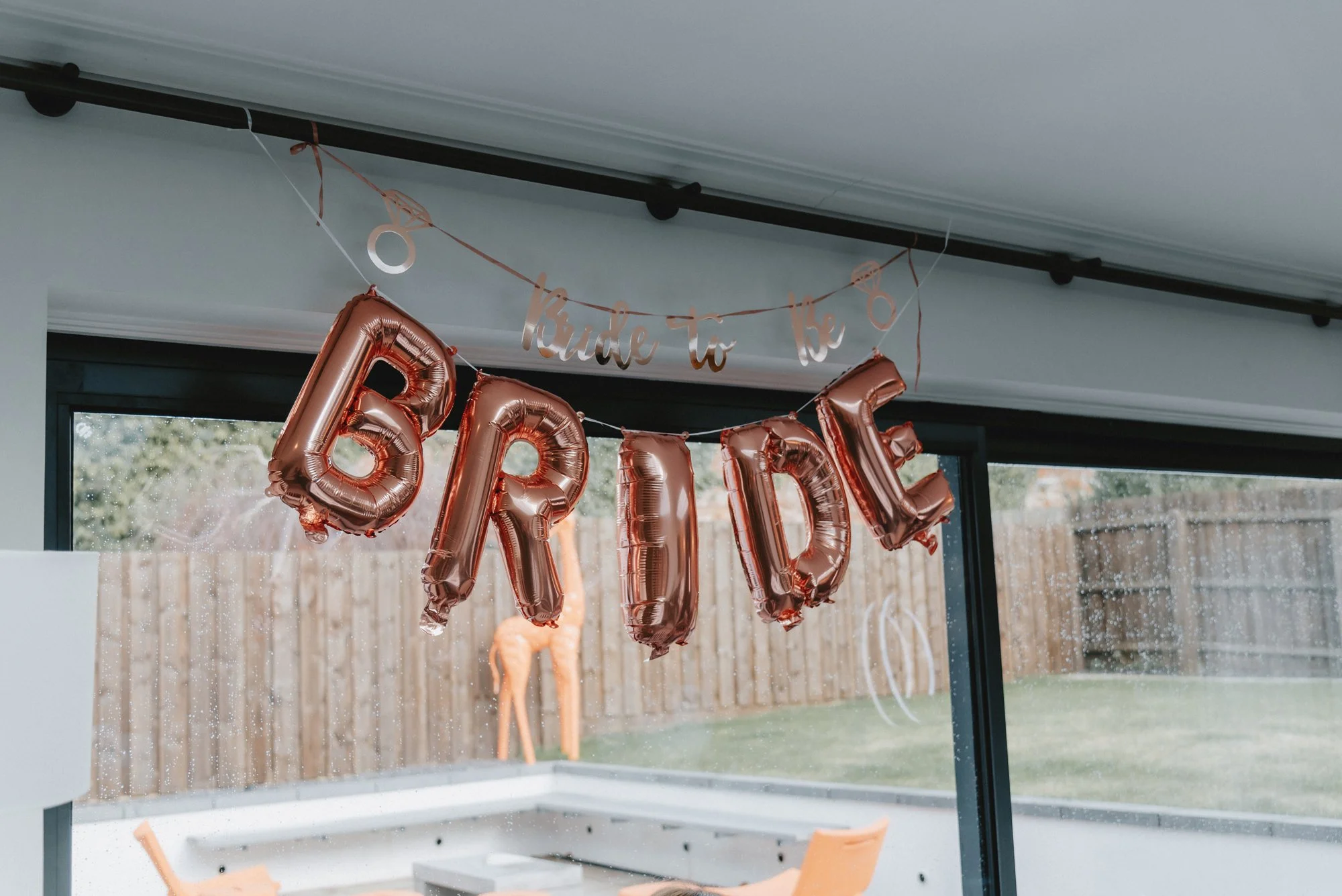 Rose gold letter balloons spelling 'BRIDE' hanging indoors with a 'Kude to be' banner above, in front of a window with a view of a wooden fence and garden.