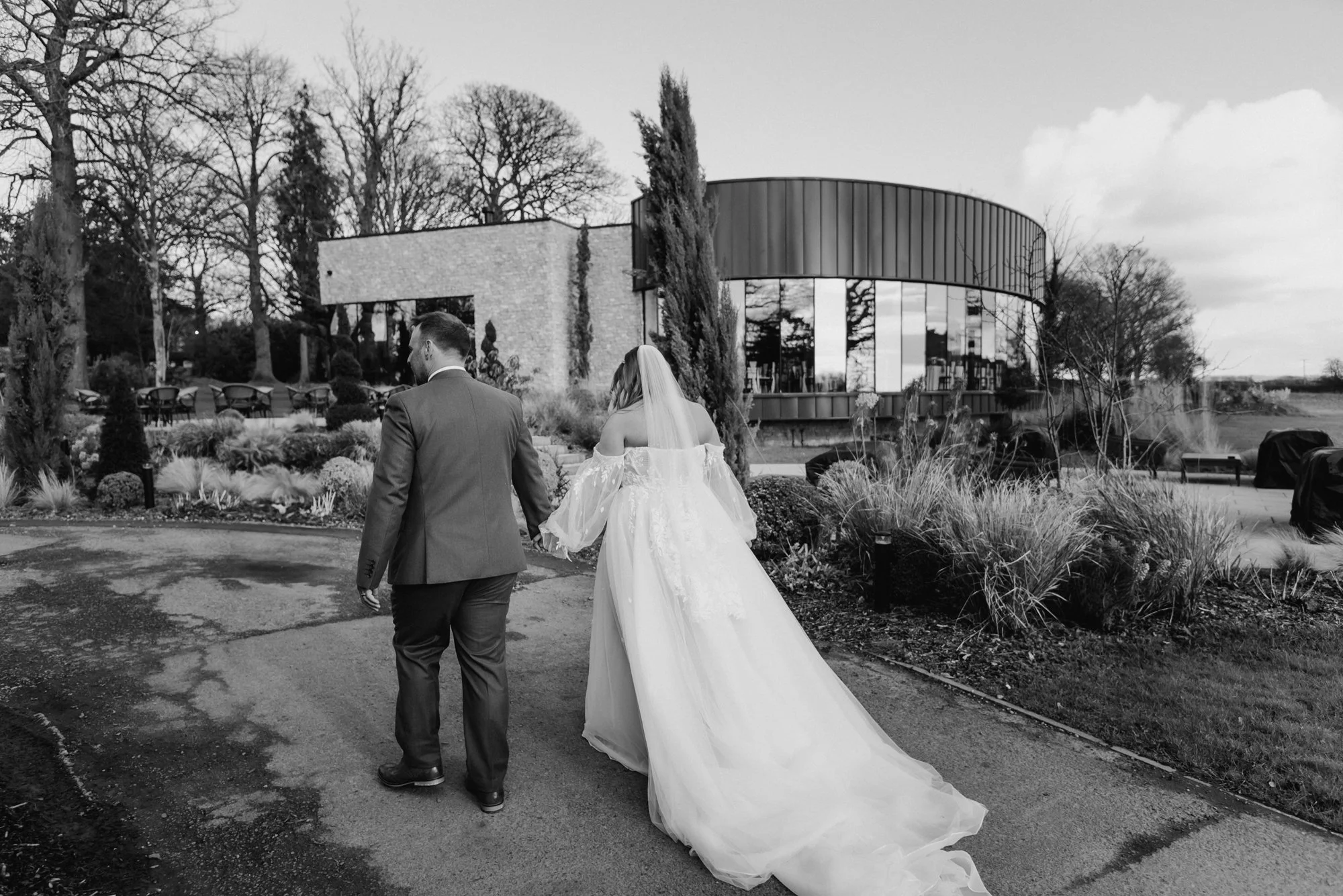 A bride and groom walk hand in hand on a paved path outside a modern building during a wedding. The bride wears a flowing wedding dress with an off-the-shoulder design and a veil; the groom wears a suit. Bare trees and landscaped bushes are in the ba
