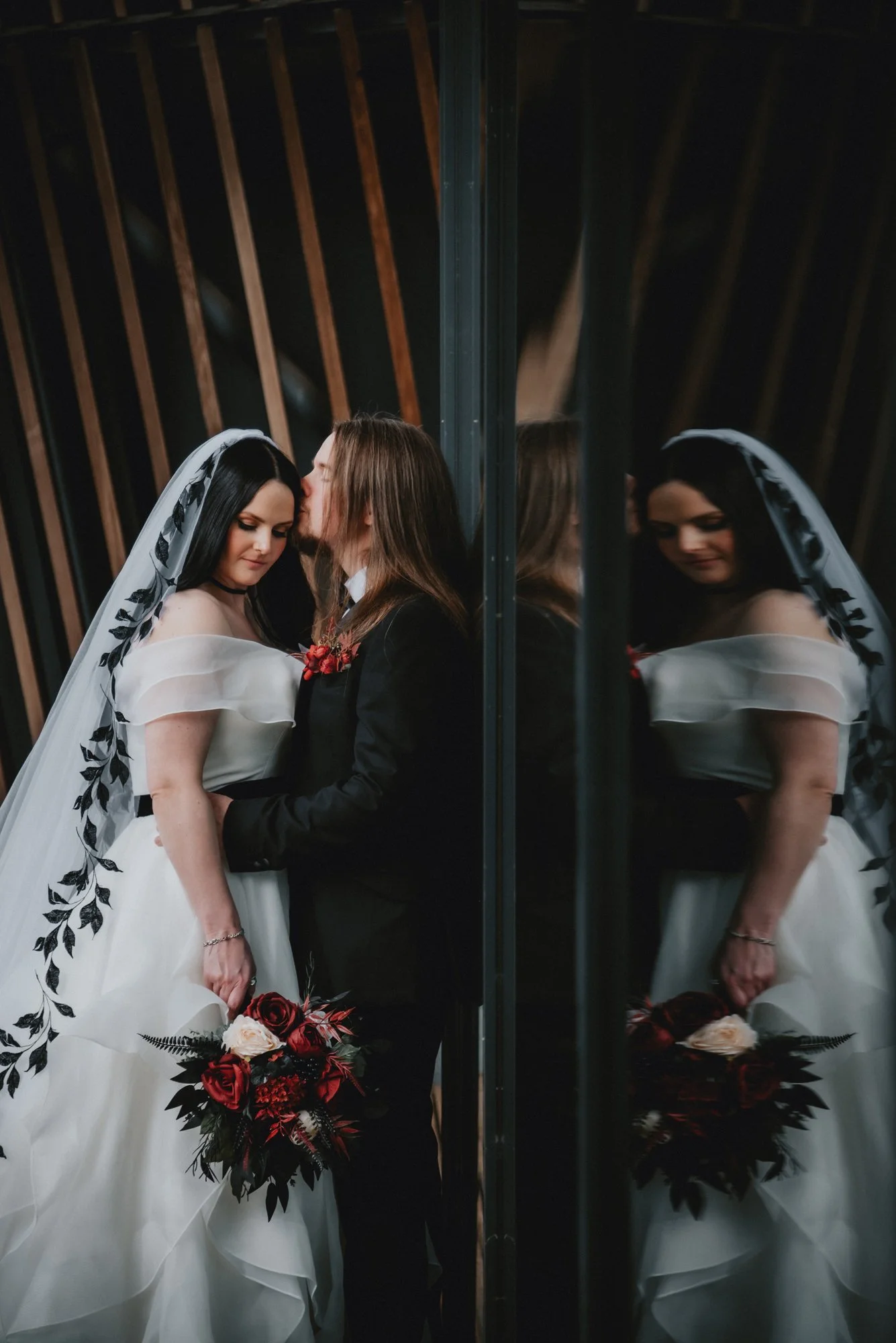 A bride and groom stand close together, with the groom kissing the bride's forehead, reflected in a mirror. The bride wears a white wedding dress with off-the-shoulder sleeves, a veil with black leaf embroidery, and holds a bouquet of red, white, and