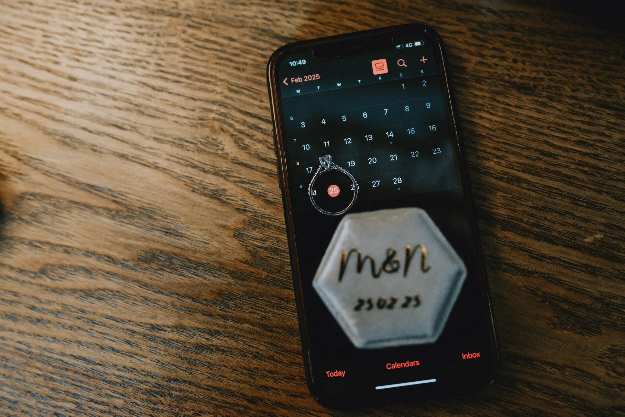 A smartphone displaying a calendar app on a wooden surface with a ring and a hexagon-shaped item with writing on it.
