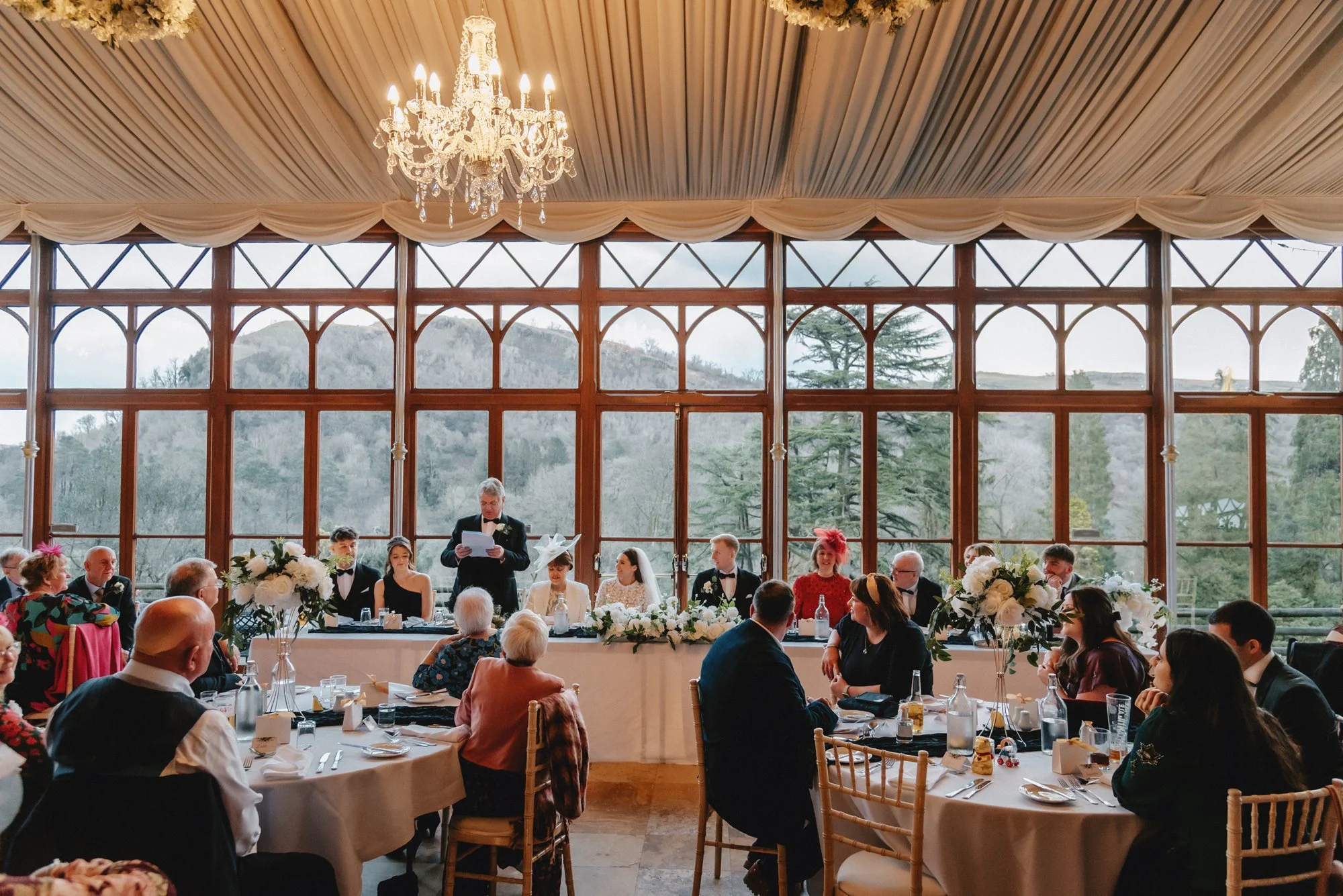 Wedding reception with guests seated at round tables, large windows revealing outdoor scenery, and a head table with the bride, groom, and family, decorated with floral arrangements, during daytime.