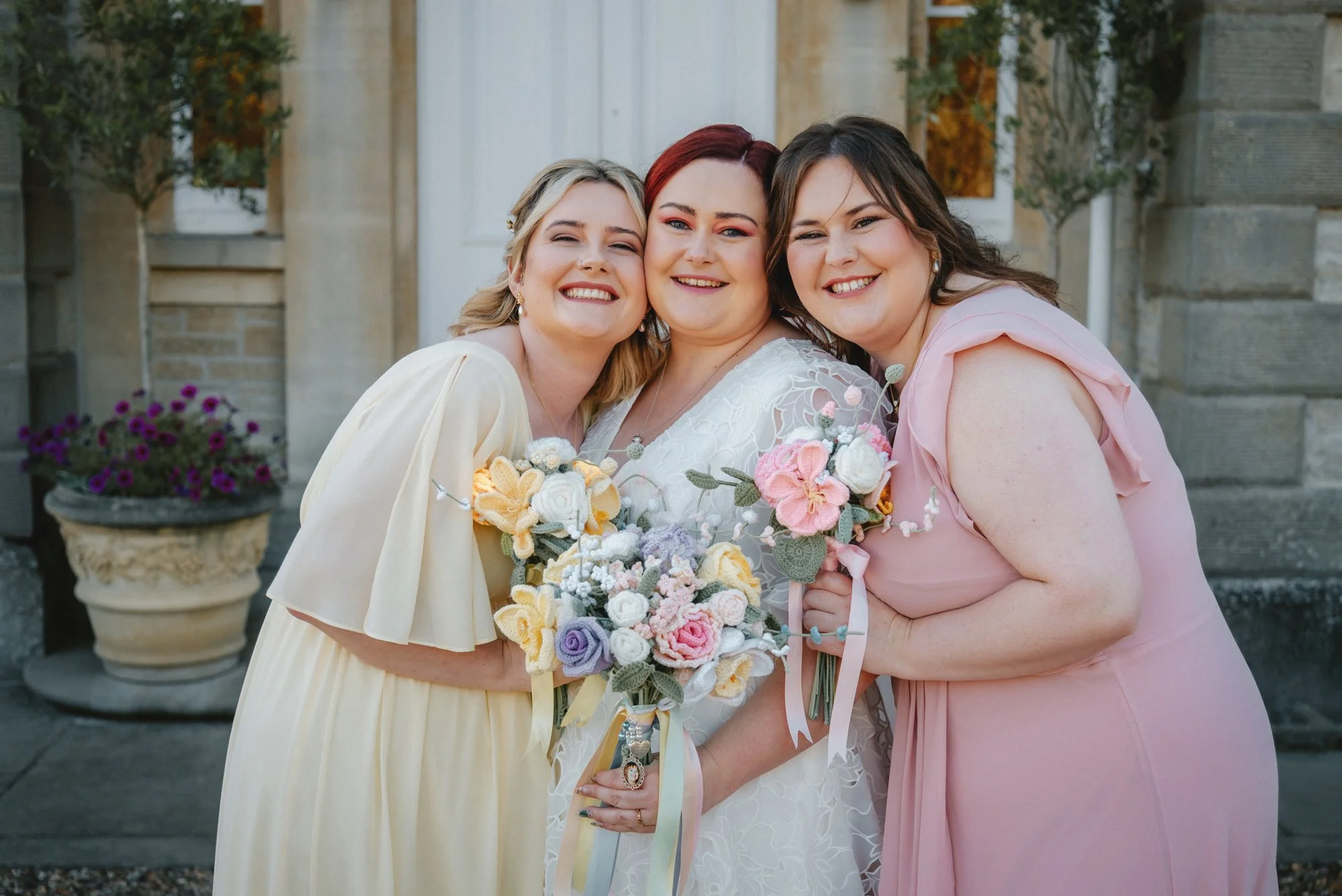 Three women smiling and hugging each other in front of a stone building, one holding a colorful bouquet of flowers, dressed in feminine attire for a celebration.