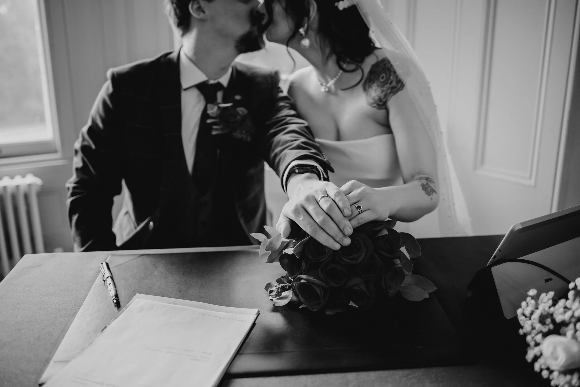 Black and white photo of a bride and groom sitting at a table, holding hands over a bouquet of roses during their wedding ceremony, with wedding rings visible on their fingers.