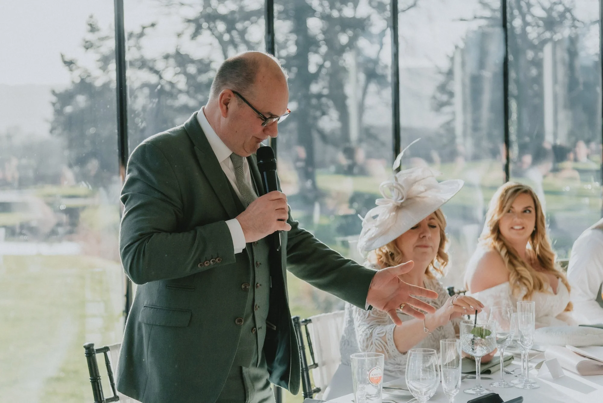 A man in a suit giving a speech at a wedding reception, with two women seated at the table, one in a large hat and the other in a white dress, during the daytime event.