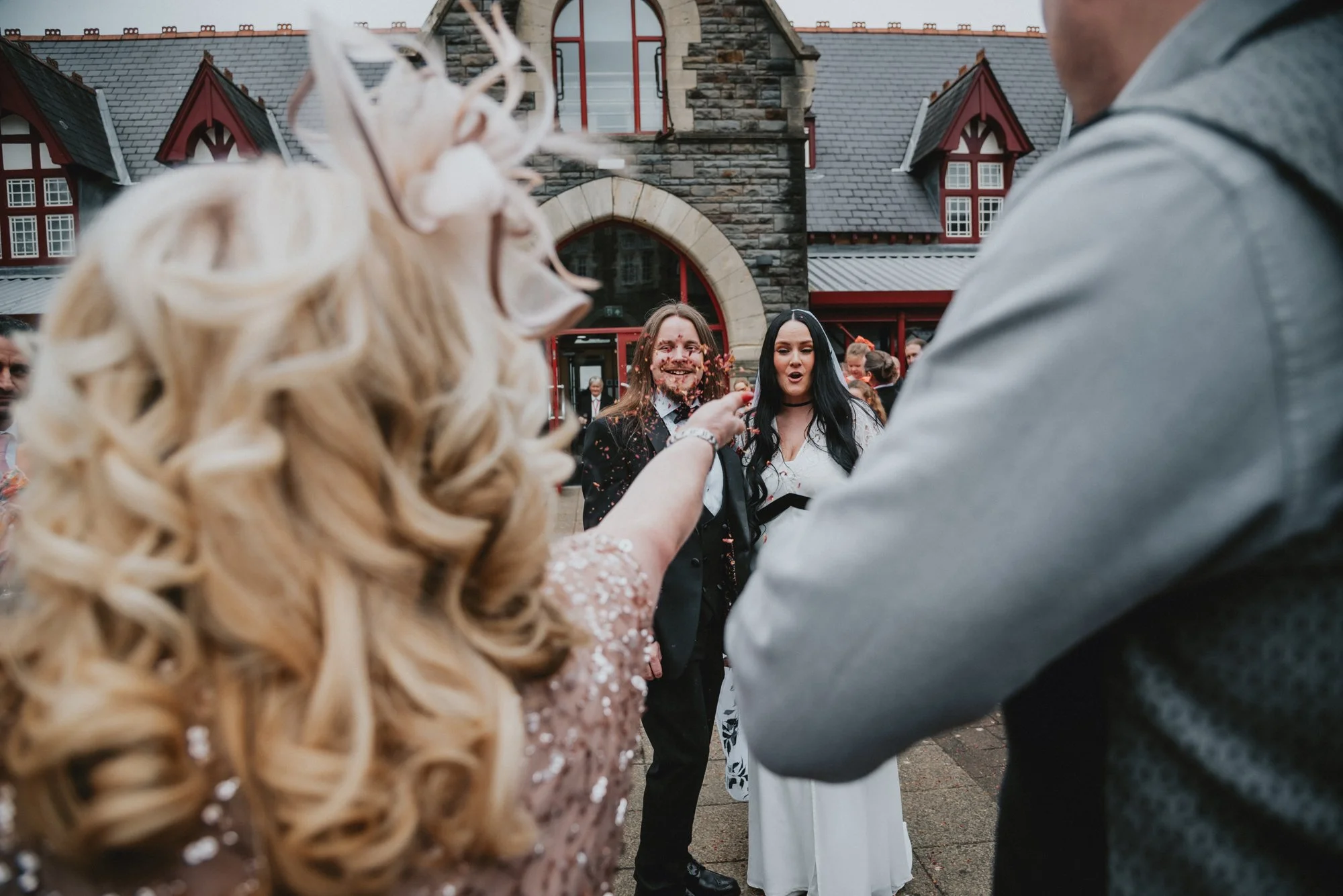 People celebrating outside a stone building with a red door and window frames, with a woman in a white dress and two men in dark suits at the center, surrounded by other guests.