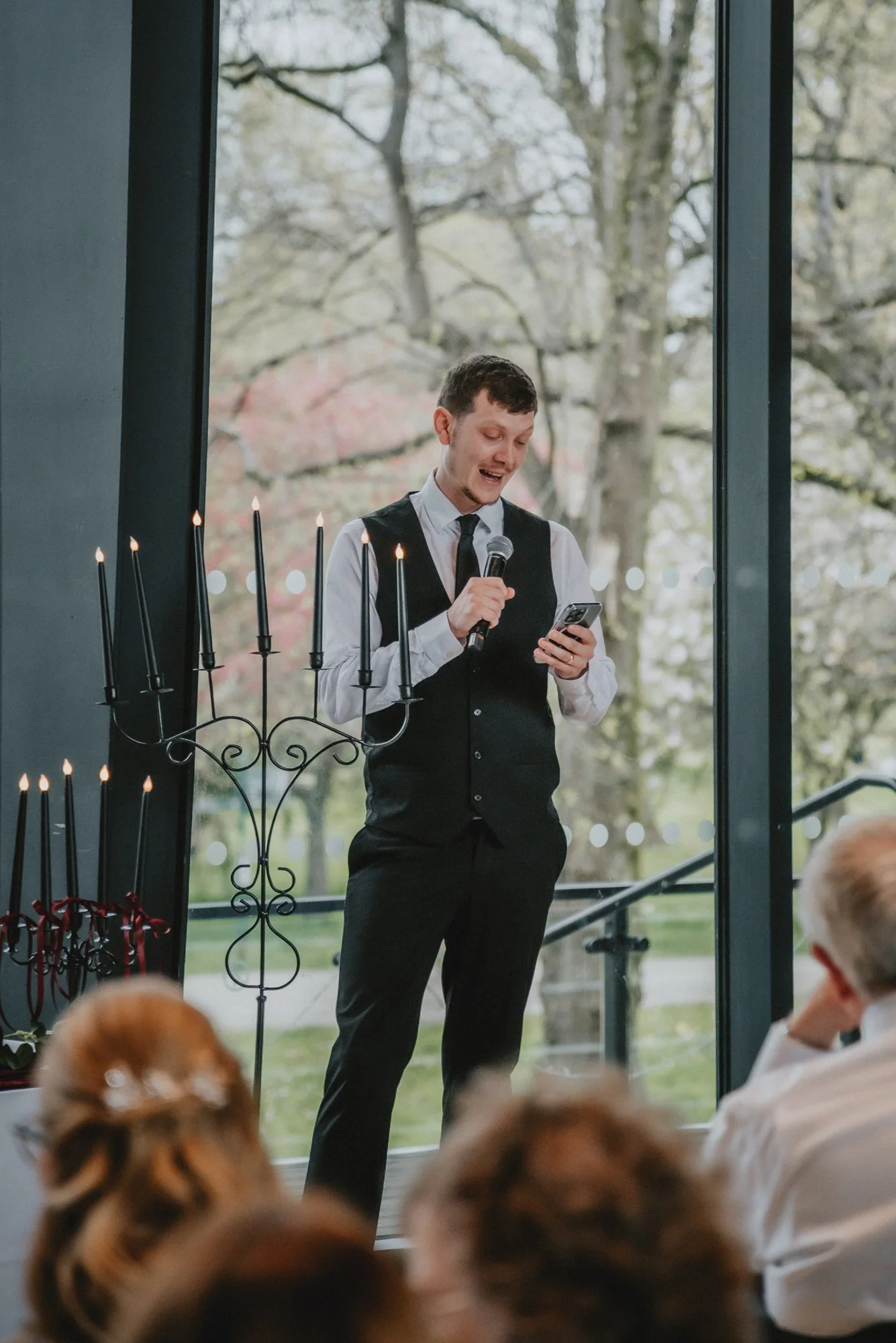 A man in a formal vest and tie speaking into a microphone while looking at his phone during an indoor event with guests watching, with large windows and trees outside.