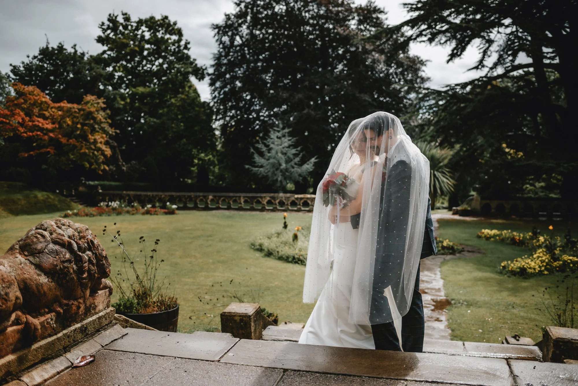 A bride and groom standing close together outdoors, with the bride holding a bouquet of flowers, both under a wedding veil, in a garden setting with trees and flowers