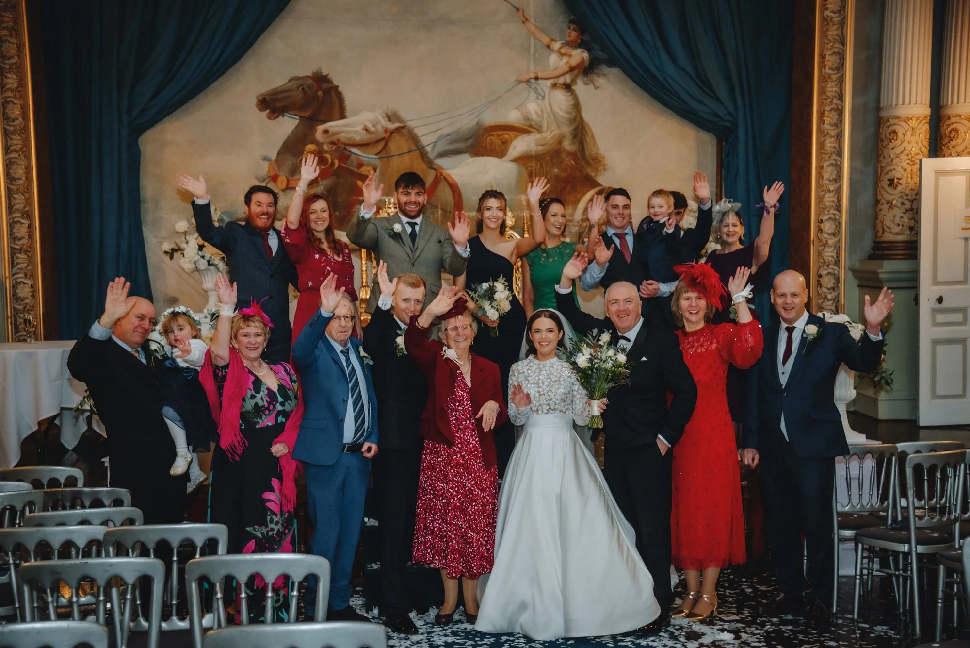 Group of people celebrating a wedding indoors, with a woman in a white wedding gown holding a bouquet, surrounded by family and friends, some wearing colorful dresses and suits, in front of a large classical mural depicting a chariot scene.