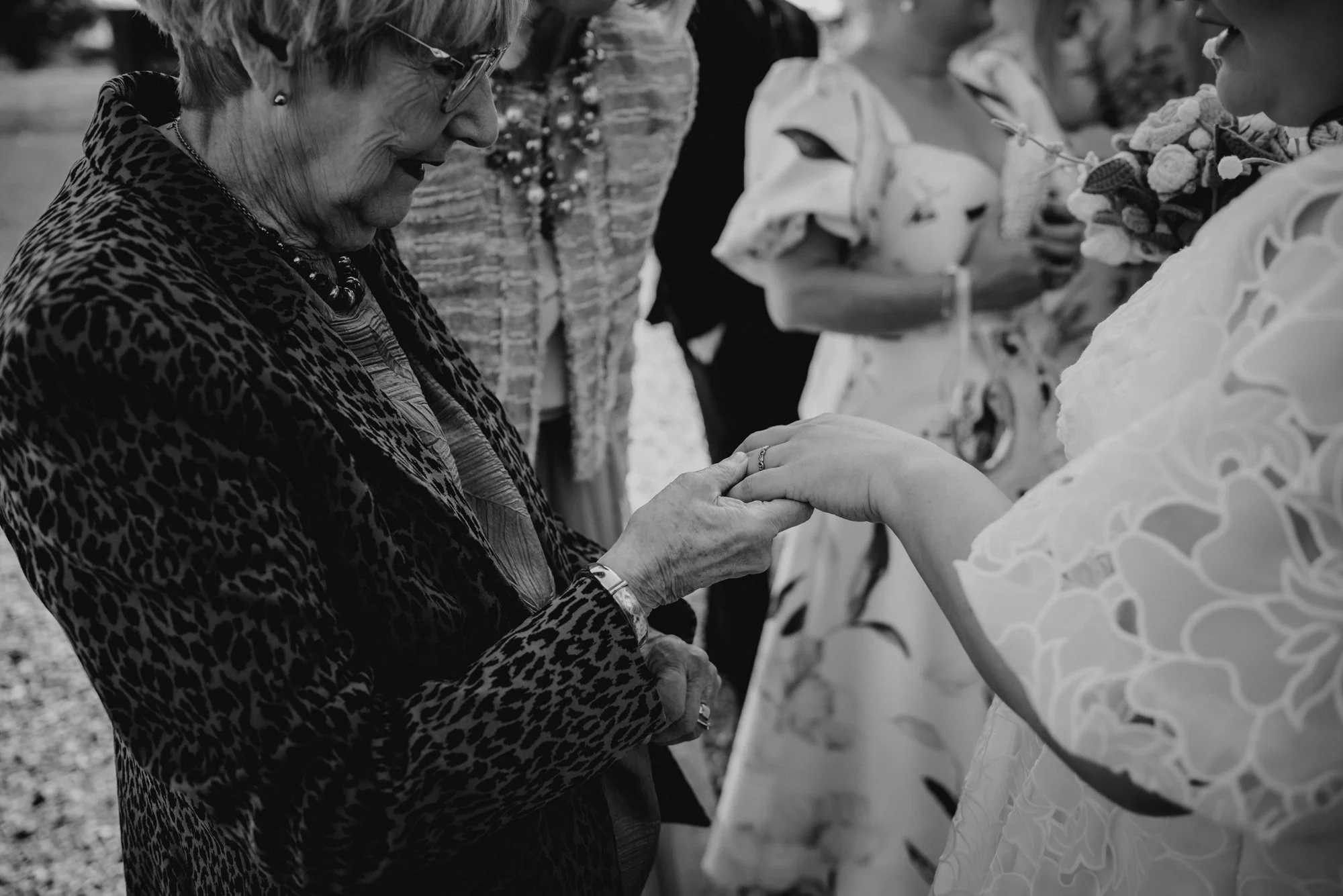 An elderly woman wearing glasses and a leopard print jacket holding hands with a woman in a white floral dress during an outdoor event.