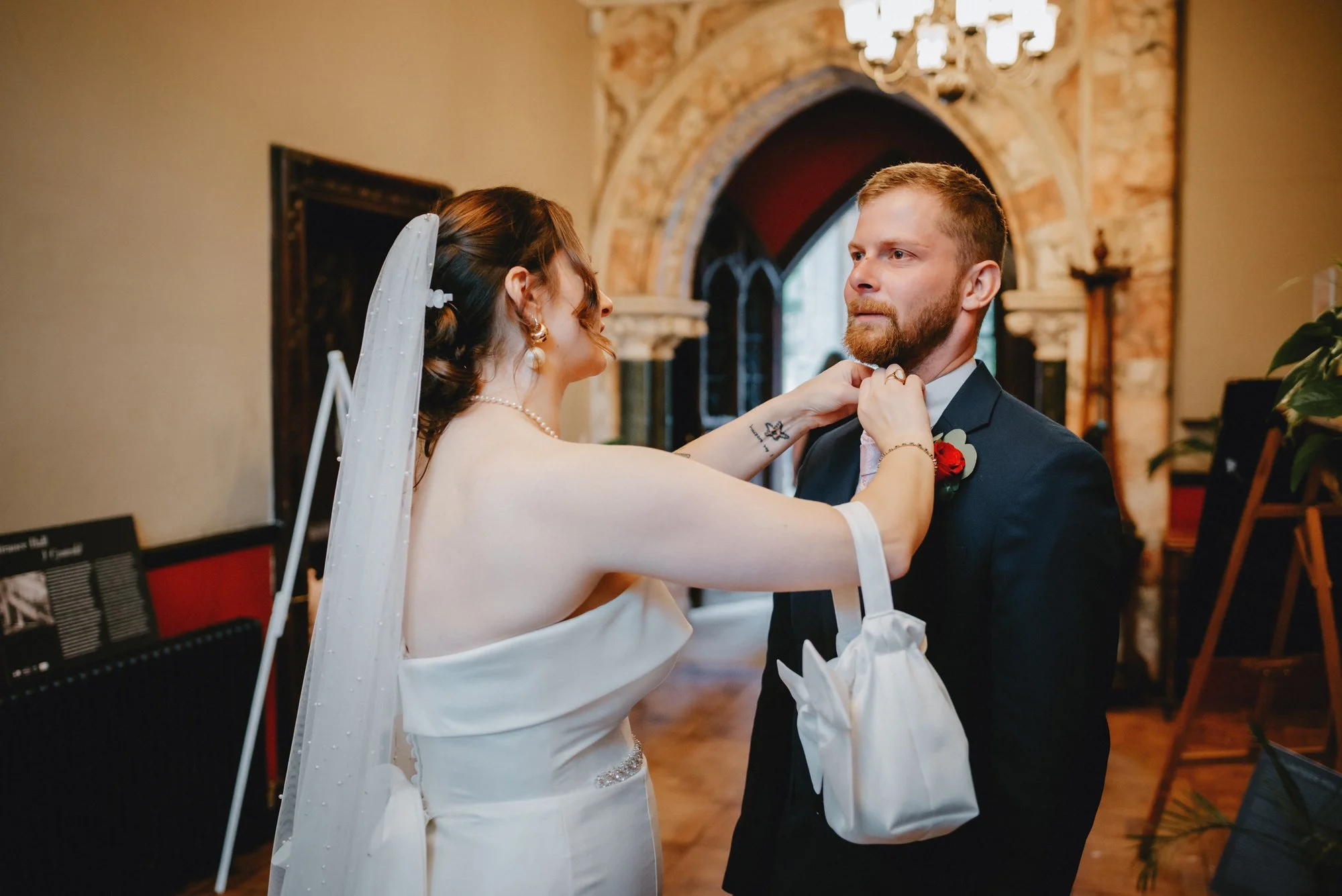 A bride adjusting a groom's tie during their wedding in a decorated church with stained glass and brick archways.