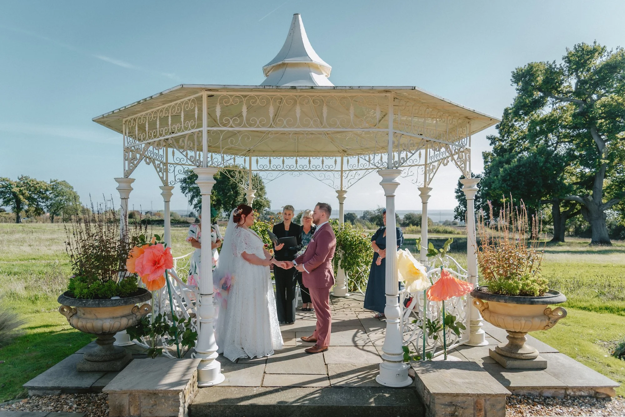 A couple getting married outdoors under a white gazebo, holding hands and facing each other, with an officiant and two women observing in the background. The setting includes large flower pots with pink and orange flowers, a sunny sky, green trees, a