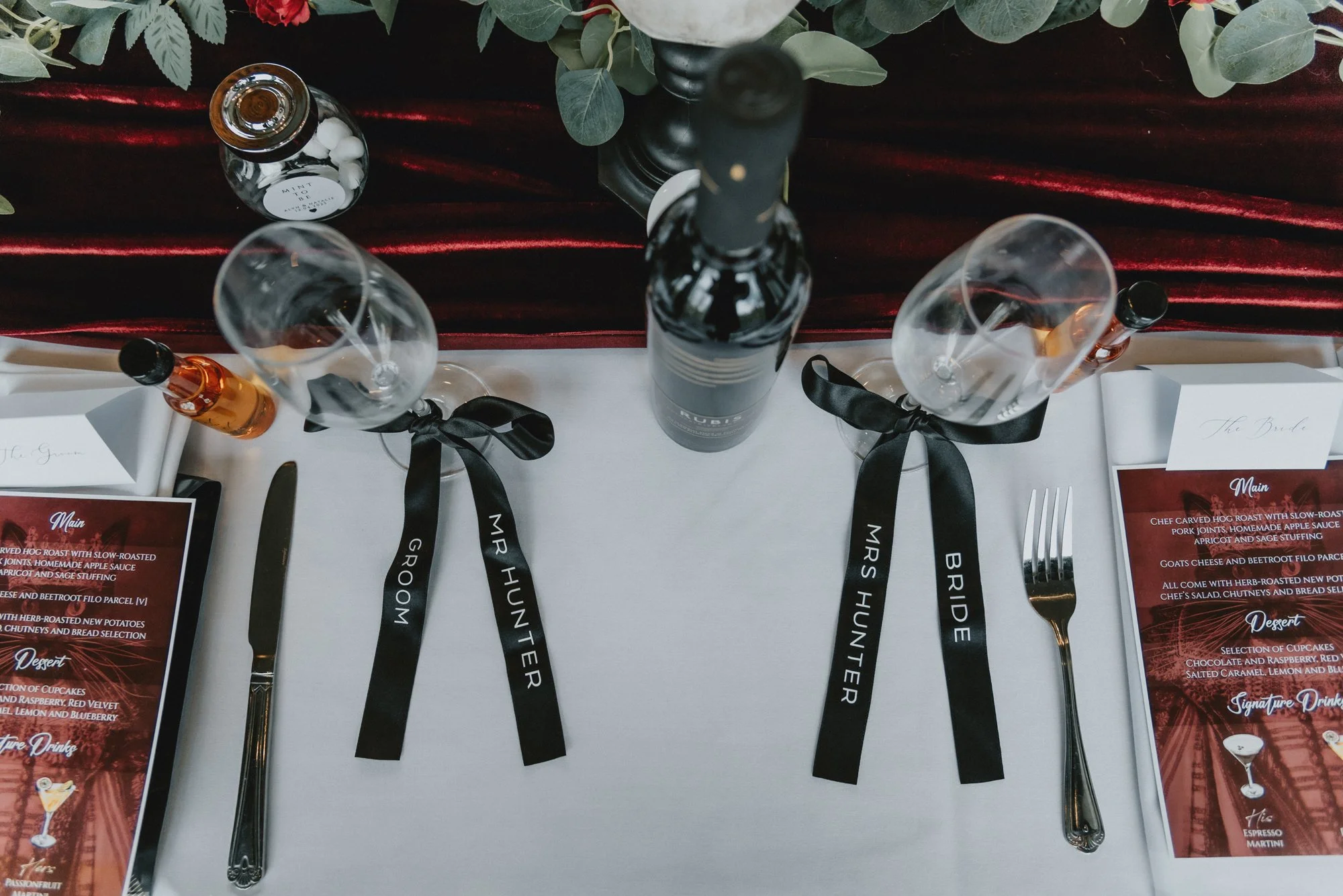 Table set for a wedding, with a white tablecloth, two wine glasses tied with black ribbons labeled "GROOM" and "BRIDE," a bottle of champagne, a small bottle of orange liqueur, a black candlestick with a black candle, a bottle of alcohol, and a weddi