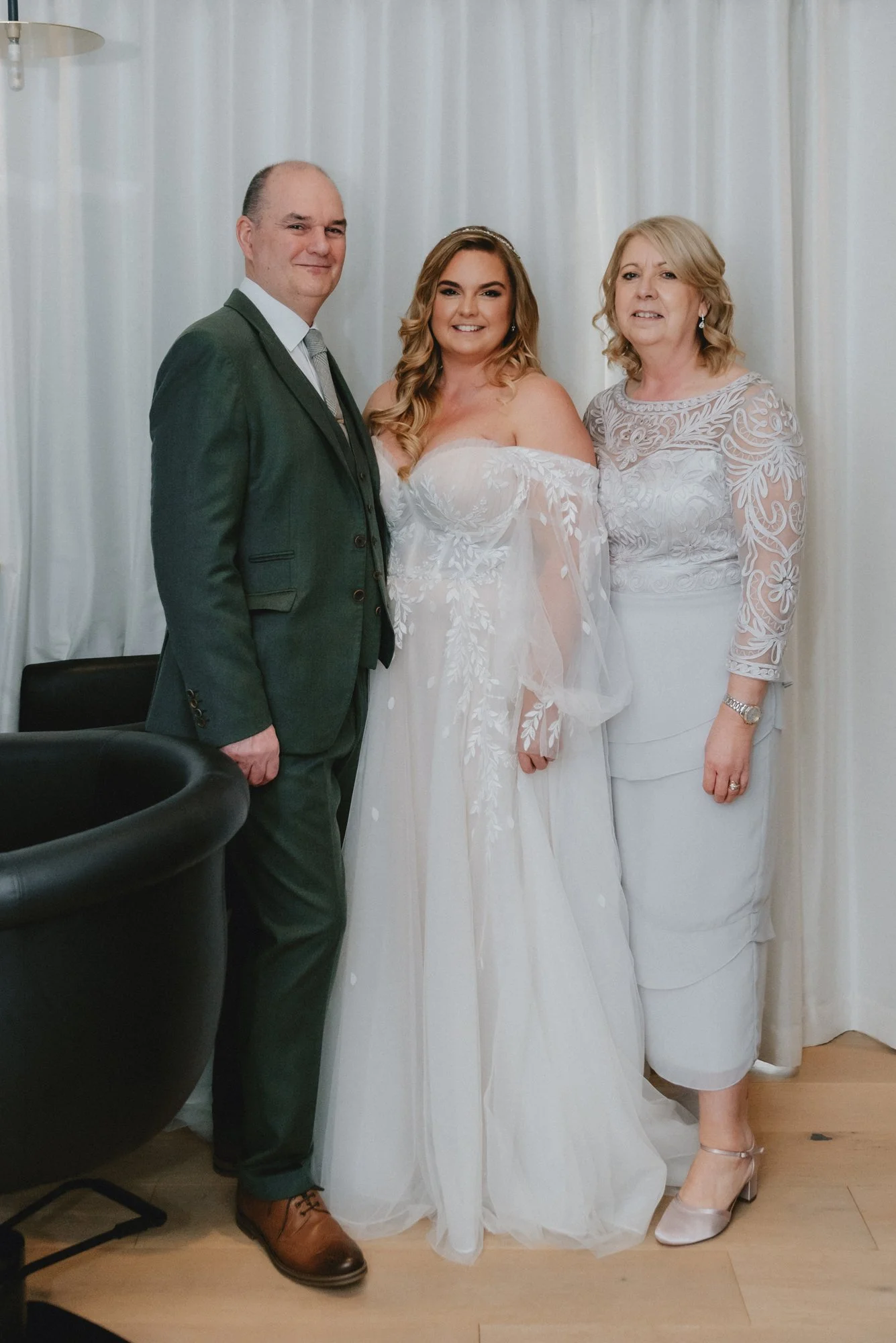A bride in a white wedding dress standing between her parents, with her father in a dark suit and her mother in a white lace dress, in front of a white curtain.