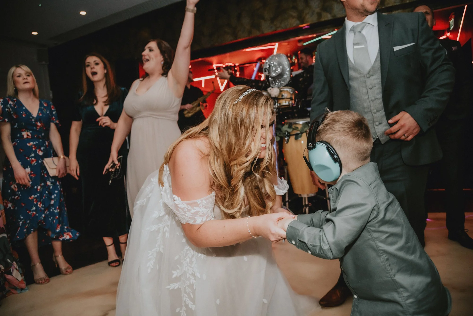 A woman in a white wedding dress is dancing and laughing with a young boy wearing large soundproof headphones. They are holding hands and appear to be enjoying themselves. In the background, several women are watching, and some are singing or cheerin