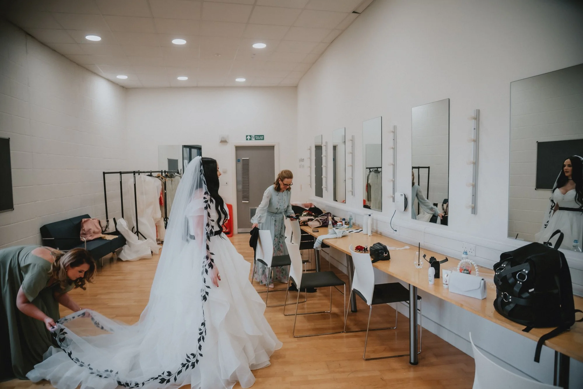 Bridal preparations with women dressing a bride in a white wedding gown in a dressing room with mirrors and makeup items.