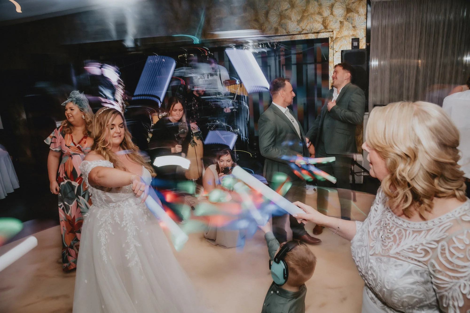 A wedding celebration with guests dancing and taking photos, including a bride in a white gown, a woman holding glow sticks, and other guests in formal attire, with colorful lights and a lively atmosphere.