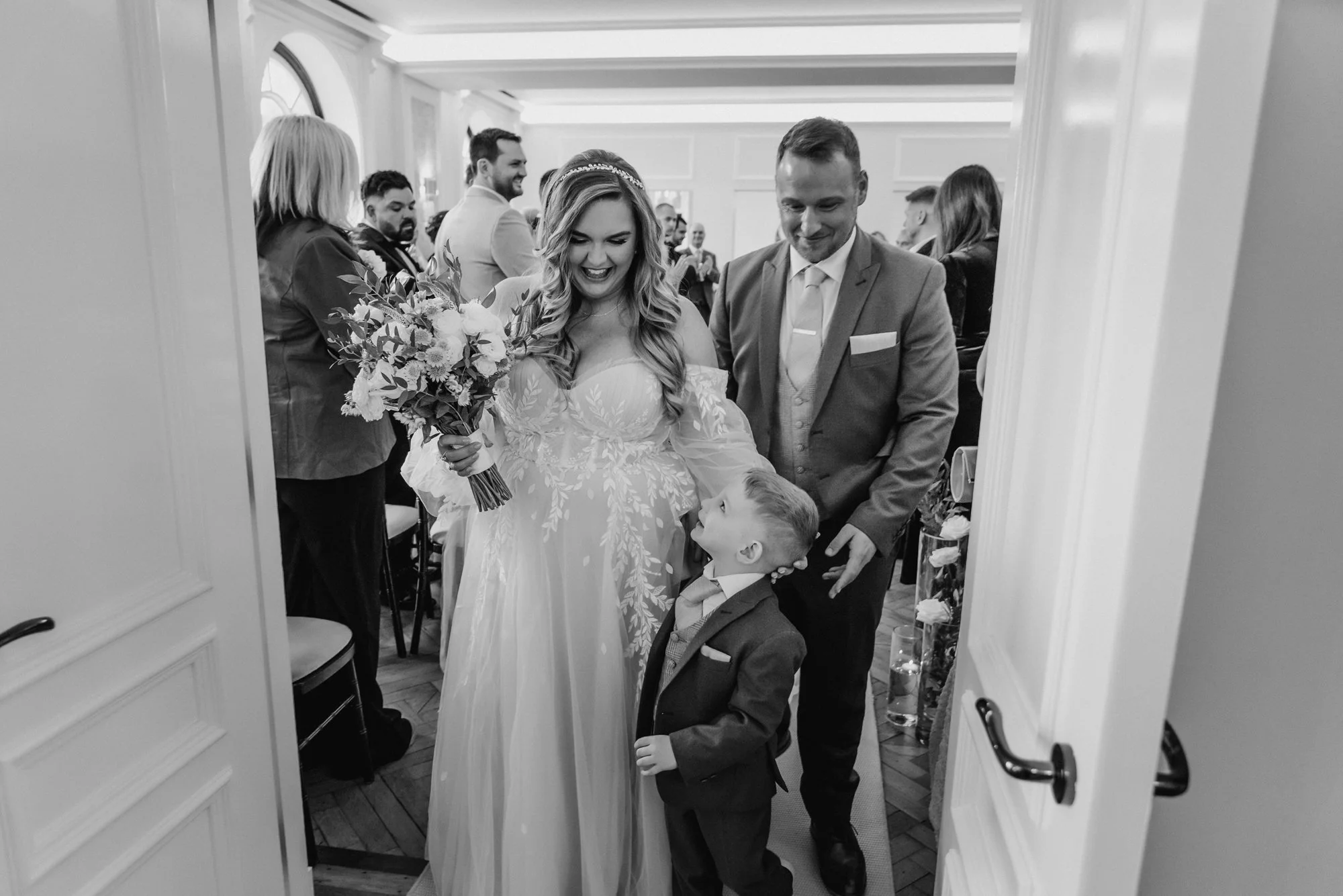 A bride in a wedding dress holding a bouquet, a young boy in a suit looking up at her, and a man in a suit standing nearby, in a room full of seated guests.