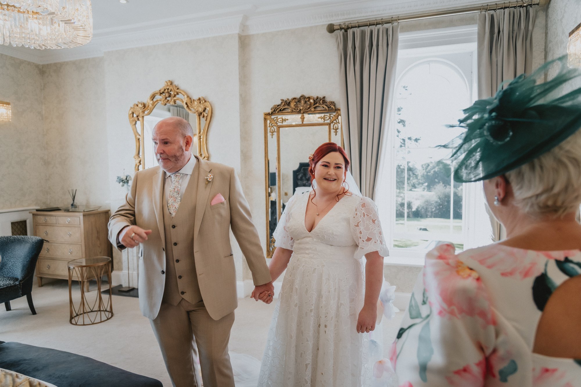 A bride holding hands with a man in a beige suit during a wedding ceremony, with a woman in floral attire and a green hat observing in a bright, elegantly decorated room.