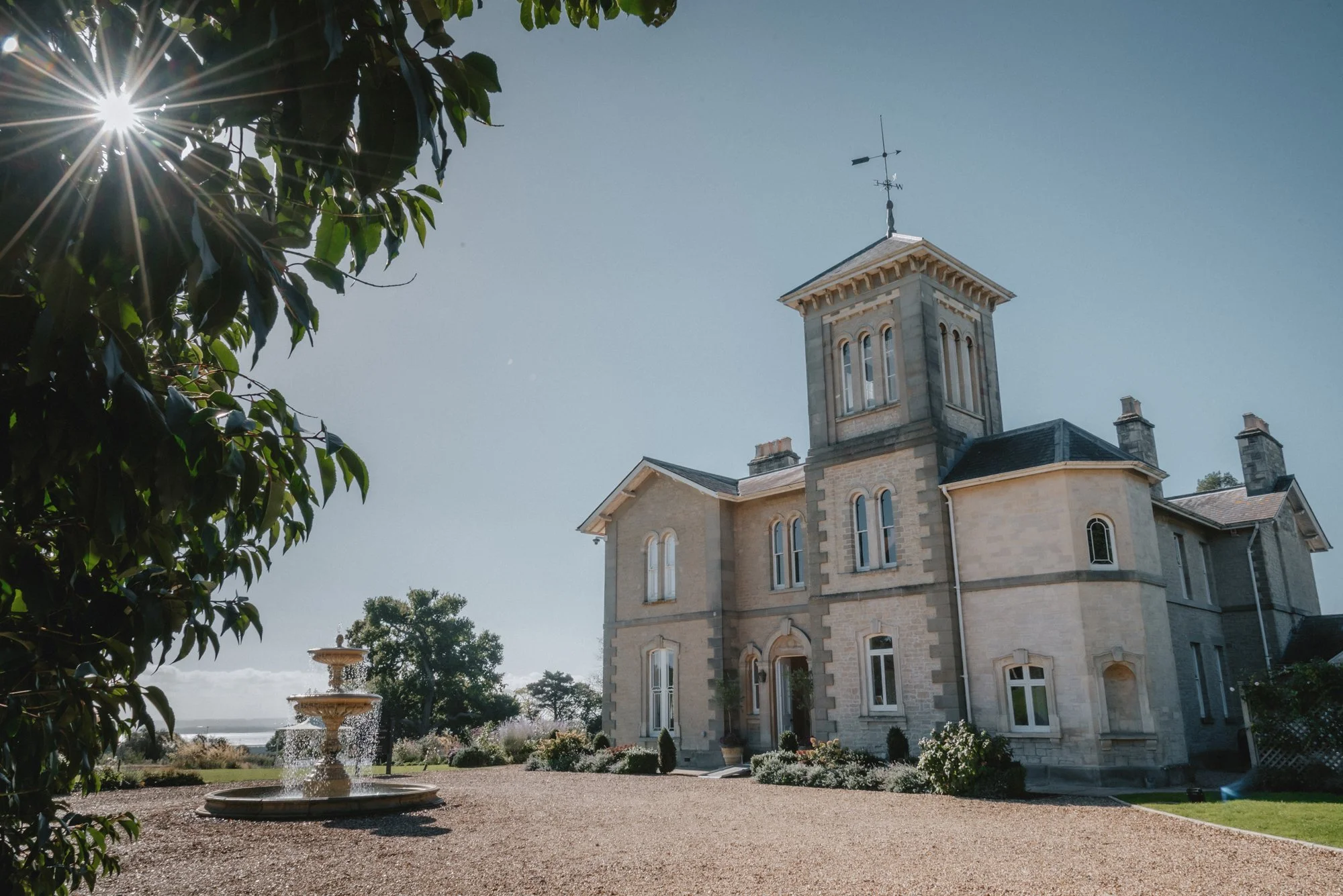 A large historic stone mansion with a tower, surrounded by a well-kept garden with flowering bushes and a gravel driveway, under a clear blue sky with the sun shining through tree leaves.