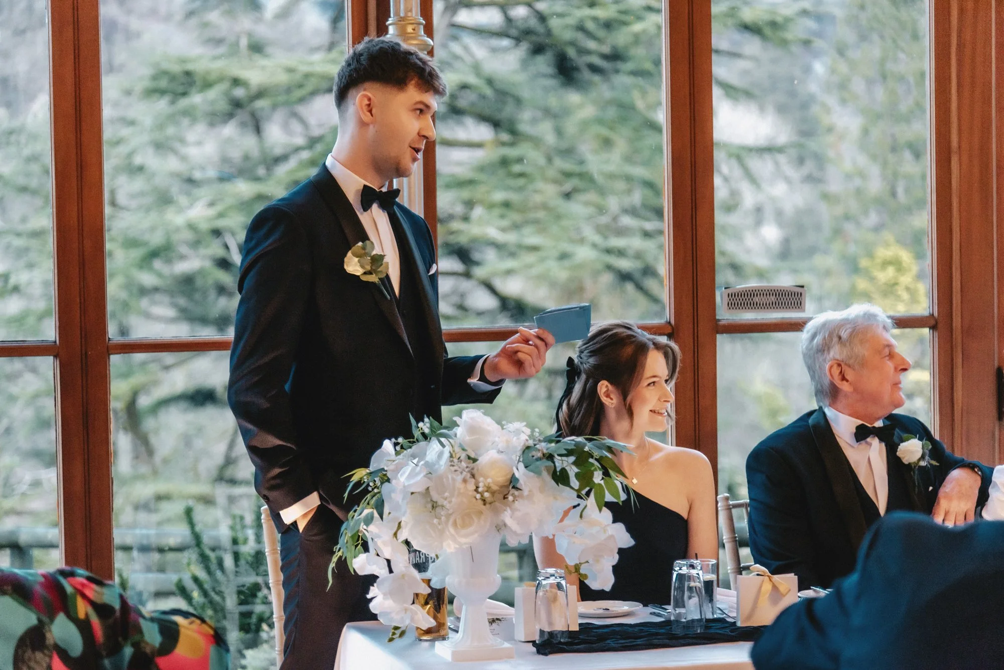 A young man in a tuxedo giving a speech at a wedding reception, standing next to a smiling woman in a black dress and an older man in a tuxedo, seated at a decorated table with a large floral centerpiece.