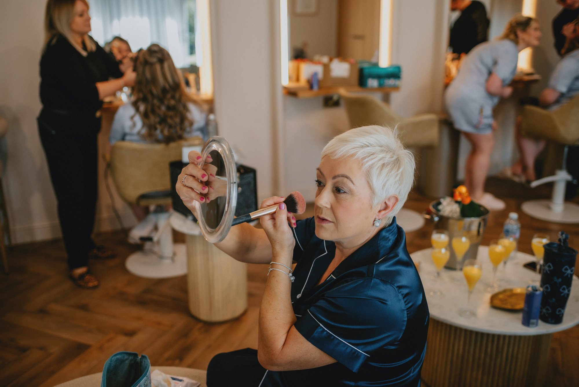 A woman with short blonde hair applying makeup using a mirror at a gathering, with other women in the background getting ready and drinks on the table.