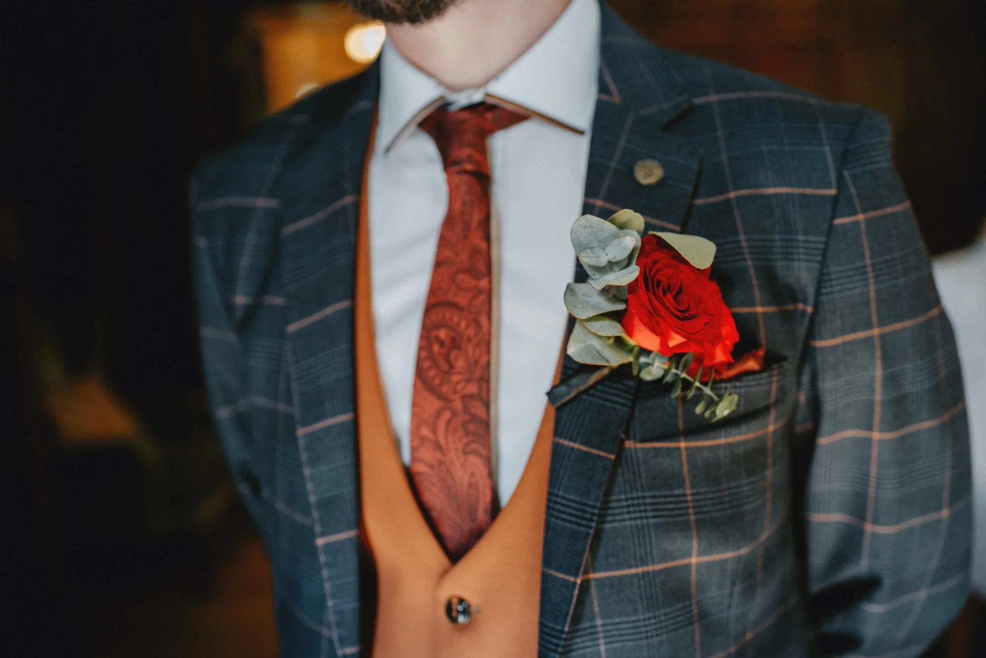 Close-up of a man wearing a dark plaid suit with a red rose boutonniere on the lapel, a white shirt, a red patterned tie, and a tan vest.