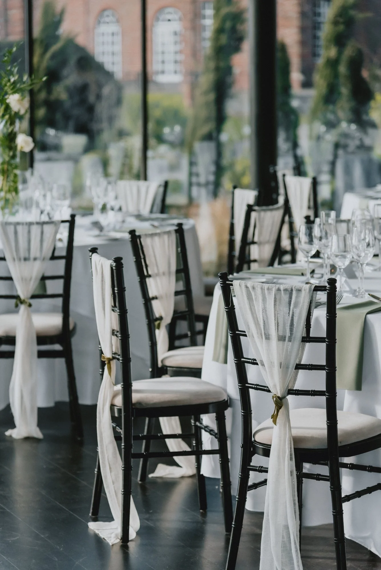 Elegant dining setup with black chairs and white draped fabric tied to the chairs, white tablecloths, and glassware, set in front of large glass windows with greenery and buildings outside.