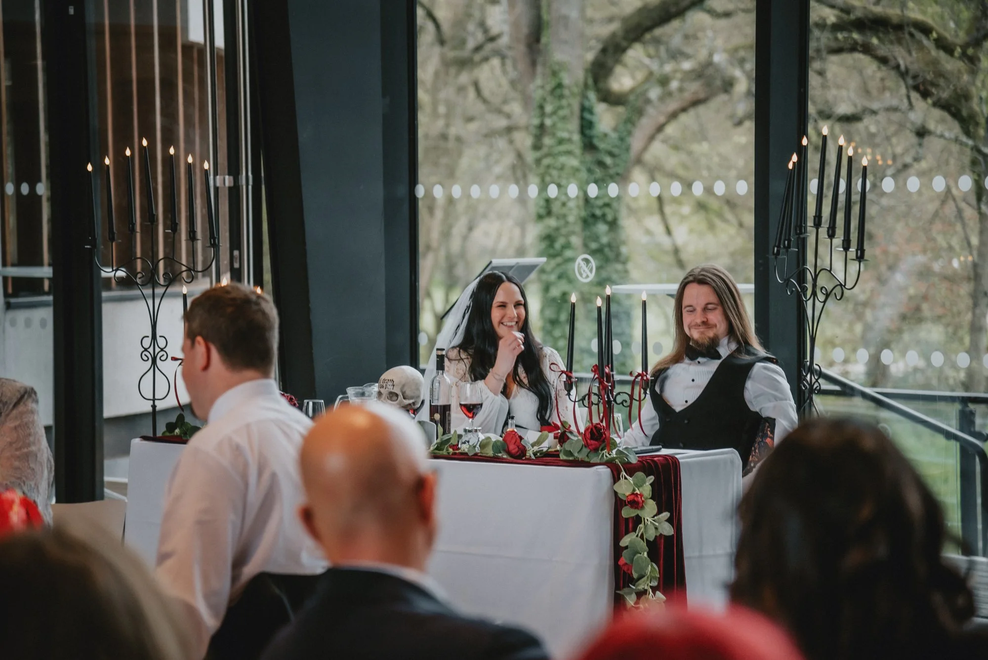 A wedding reception with the bride and groom sitting at a decorated table, smiling and enjoying the moment, with guests seated in front. The decor includes black candles with red ribbons, green foliage, and a skull. Large windows and candle-style wal