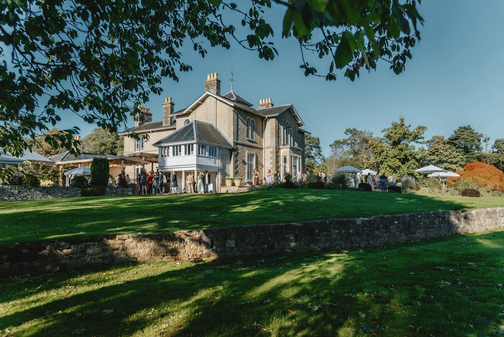 A large historic house with many windows and chimneys, surrounded by a lush green lawn and trees, with people gathered outside for an event under umbrellas.